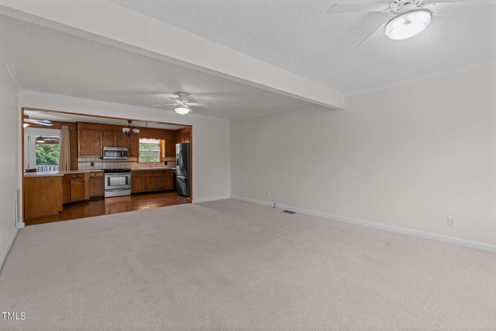 A spacious, unfurnished living room with beige carpet and white walls, connected to a kitchen with wooden cabinets and stainless steel appliances. Ceiling fans with lights are present in both areas. Natural light filters through the windows.