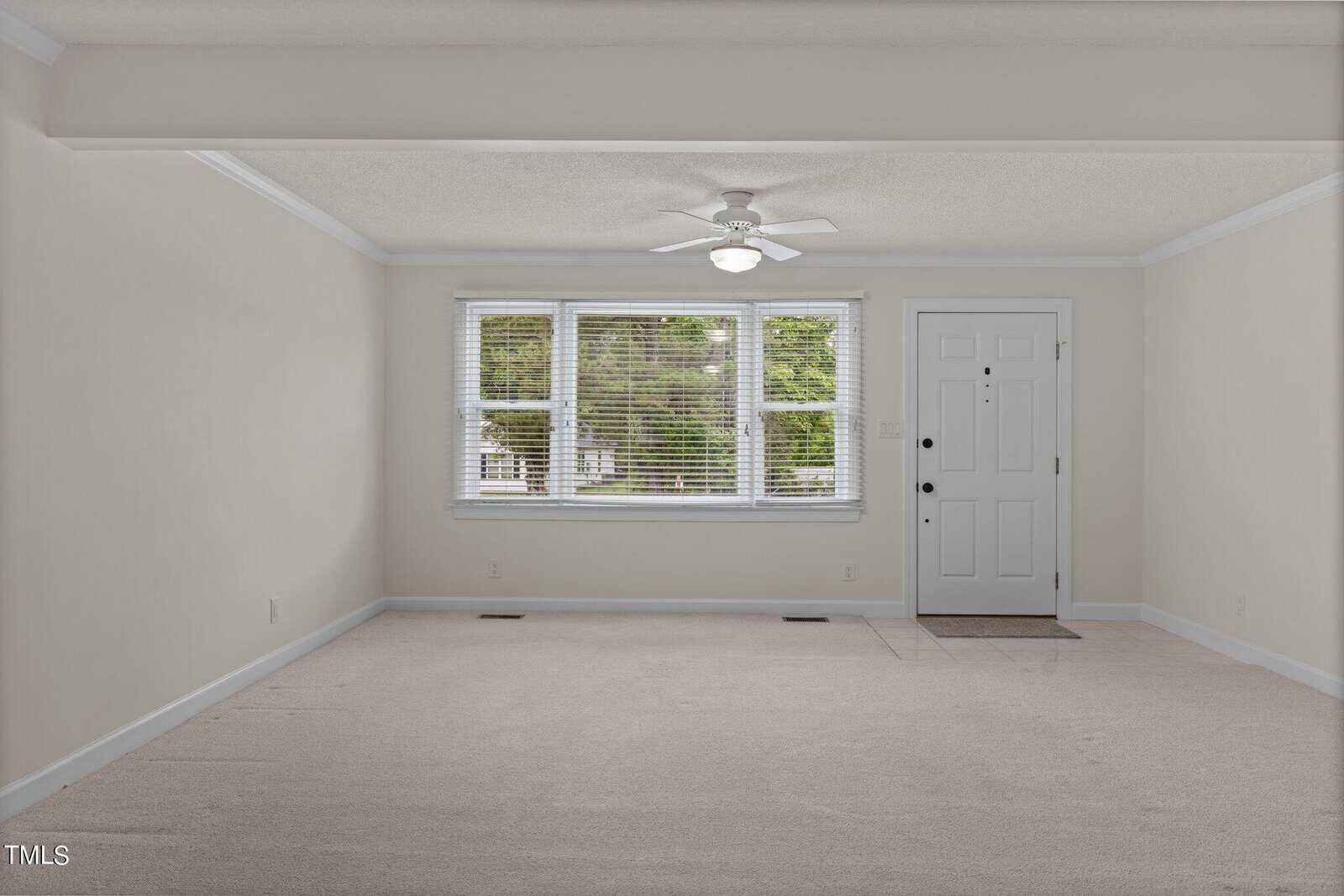An empty, carpeted room with off-white walls, a ceiling fan with light, and a large window with blinds partially open. A white door with a small window is on the right side, and the space is well-lit by natural light.