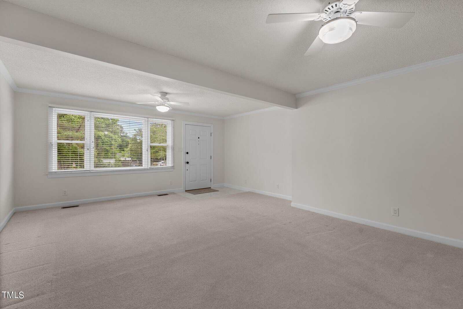 A spacious, empty room with light-colored walls and carpet, featuring two ceiling fans with lights. A large window and a white door are visible at the far end of the room, allowing natural light to enter.