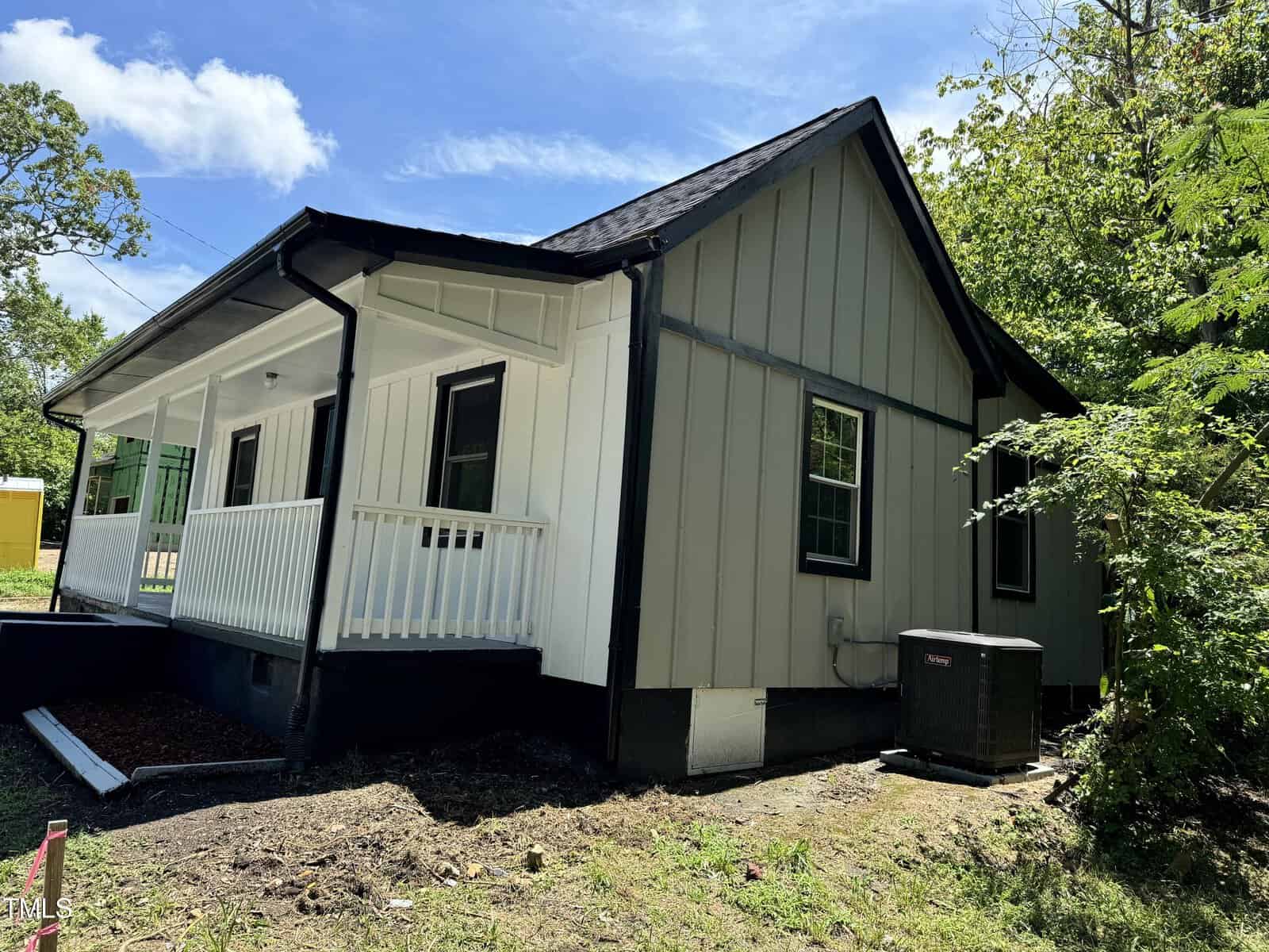 A small house with white and gray siding, black roof, and porch railings under a clear blue sky. Surrounded by trees and greenery, with an air conditioning unit on the side. Shadows indicate midday sunlight.