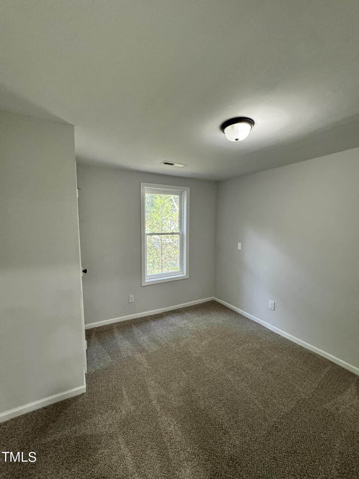 A small empty room with gray walls and a beige carpet. There is a single window with a view of greenery outside. A ceiling light fixture is visible, and a partially open door is on the left side of the image.