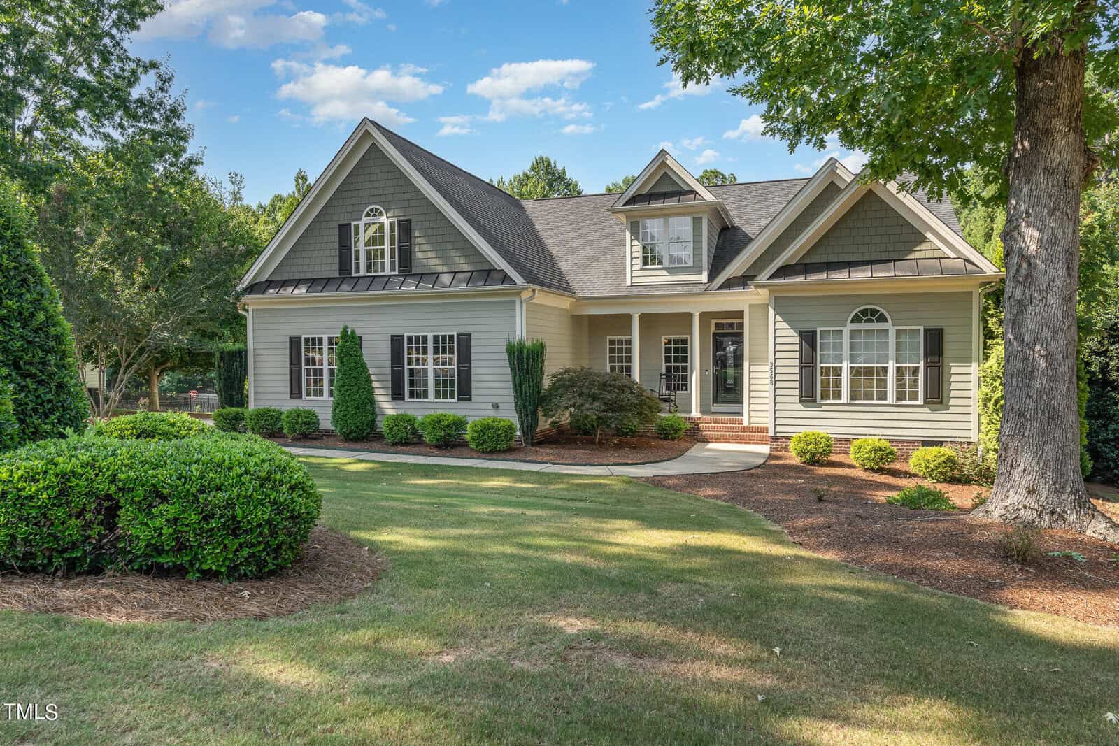 Nestled at 3568 Overlook Court in Wake Forest, this charming two-story house features a green facade, large windows, and a welcoming front porch. It's surrounded by a well-manicured lawn, lush bushes, and stately trees under the clear blue sky.