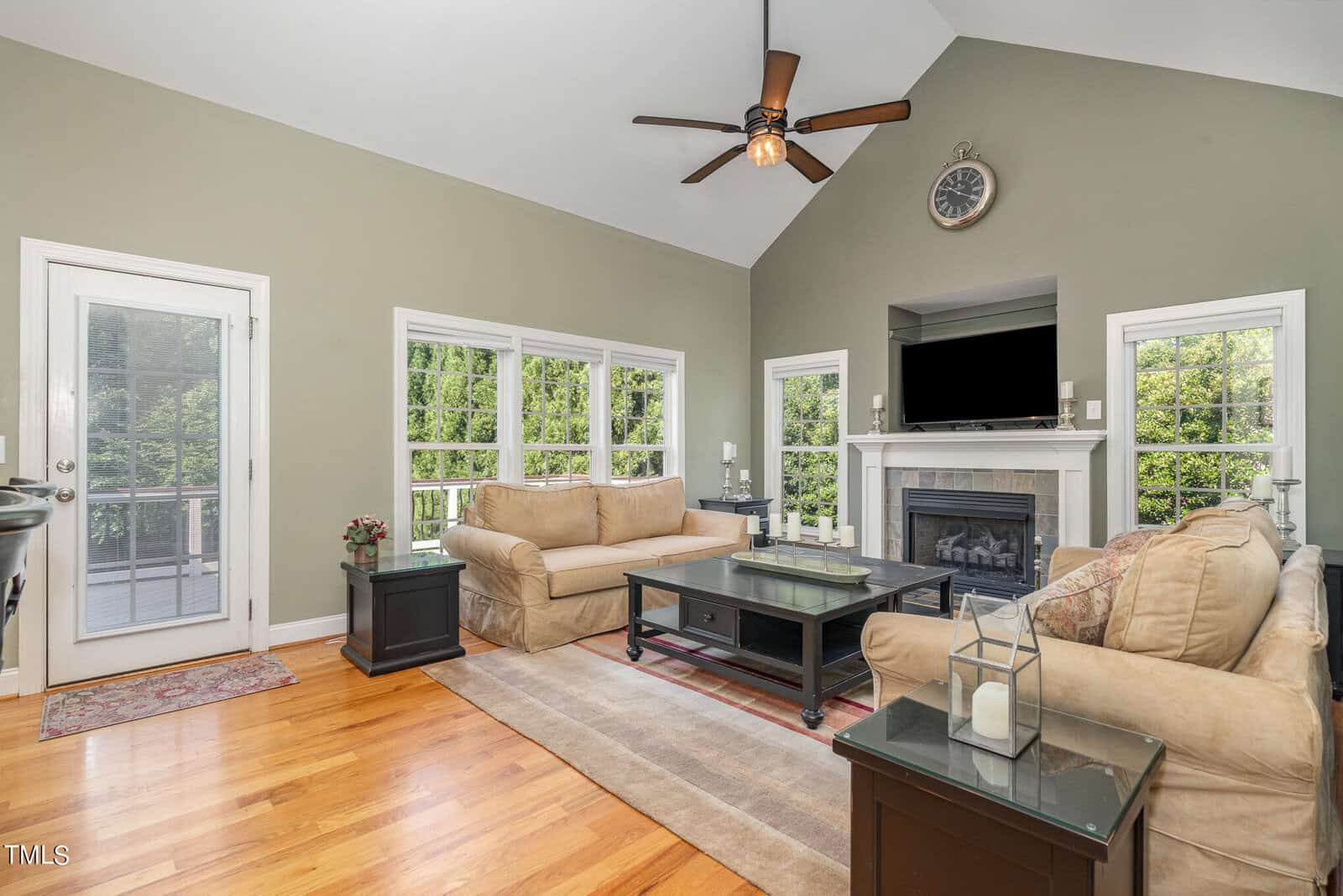 A spacious living room with two beige sofas, a fireplace, and a wall-mounted TV. Large windows let in natural light, and a ceiling fan hangs from the vaulted ceiling. French doors lead to an outdoor area. The room has hardwood floors and tasteful decor.