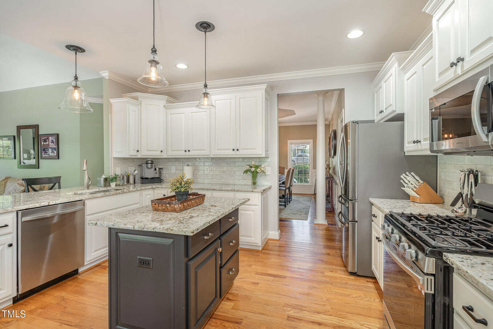 Modern kitchen with white cabinets, stainless steel appliances, and a central island with granite countertops. Pendant lights hang above the island. Hardwood floors complement the light green wall and decor accents.