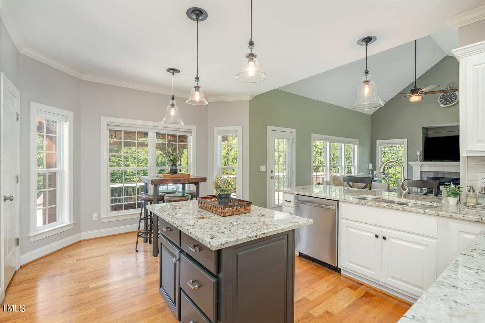 Bright kitchen with hardwood floors, featuring an island with a dark base and granite countertops. Pendant lights hang from the ceiling. Large windows provide natural light, and there's a door leading to an outdoor area. White cabinetry lines the walls.