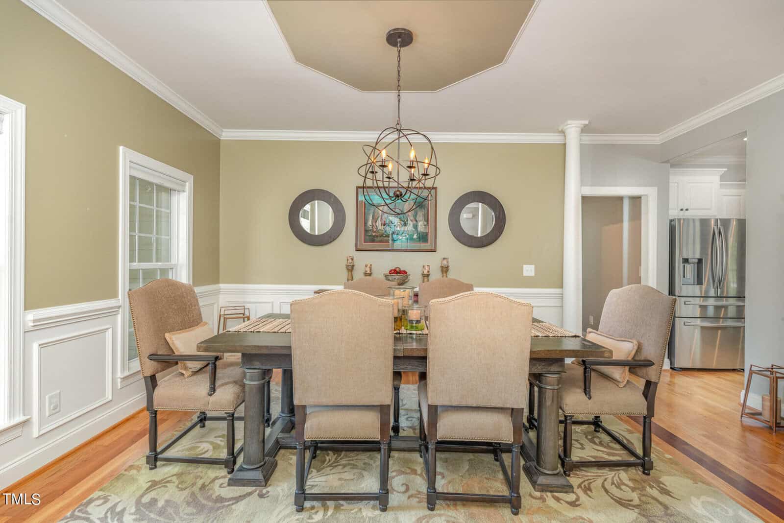A dining room with a dark wooden table surrounded by six beige upholstered chairs. A round chandelier hangs from the ceiling. The walls are light green with white wainscoting. Two round mirrors and a painting adorn the back wall.