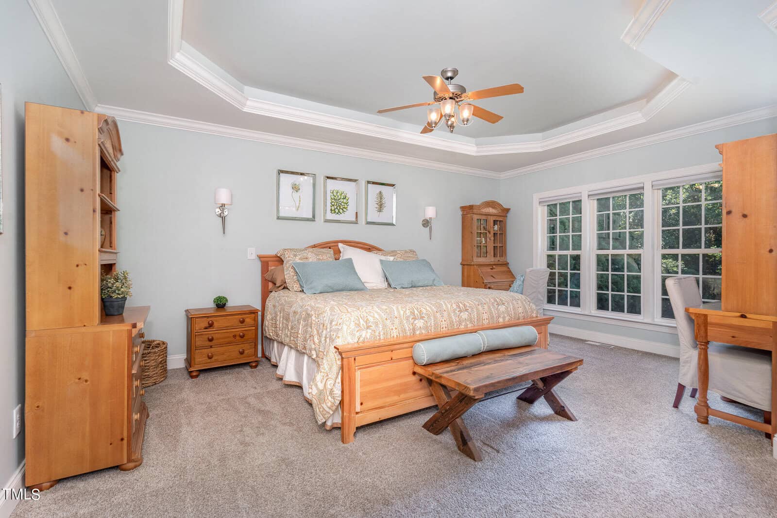 A spacious, elegantly designed bedroom with a wooden bed, matching nightstands, and a bench. The room features a tray ceiling with a ceiling fan, a large window letting in natural light, and neutral-toned decor for a relaxing atmosphere.