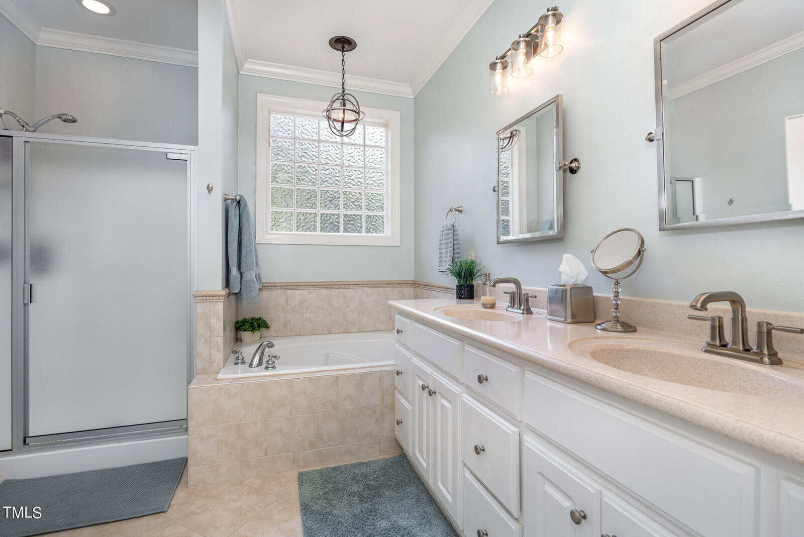 A modern bathroom with a shower, bathtub, and a double sink vanity. The room features white cabinets, a large mirror, and two light fixtures. A small window lets in natural light, and a gray towel hangs near the bathtub.