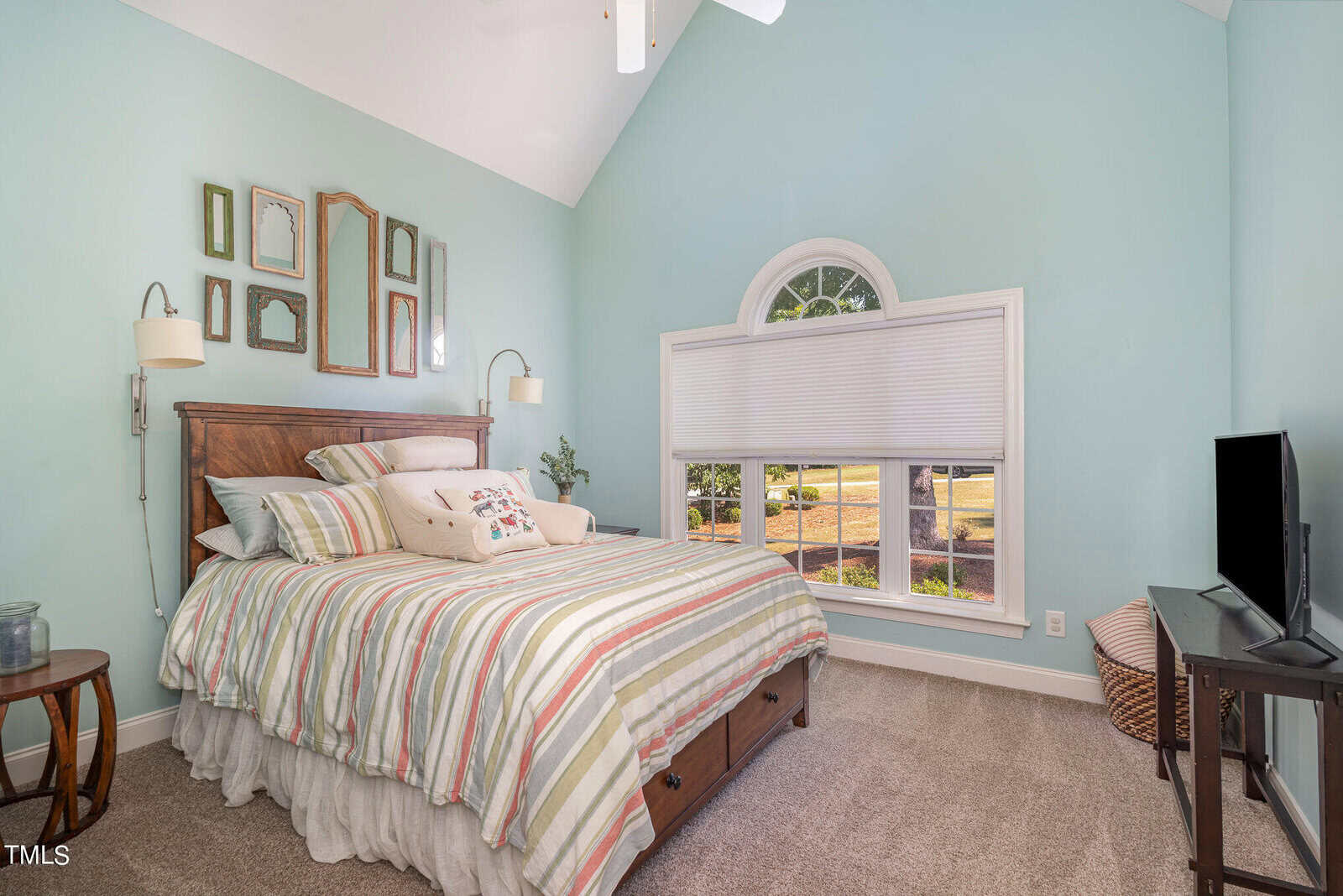 A cozy bedroom with a tall, vaulted ceiling features a bed with a striped comforter, decorative pillows, and a wood headboard. Mirrors adorn the wall above the bed. A large arched window lets in natural light, and a flat-screen TV is in the corner.