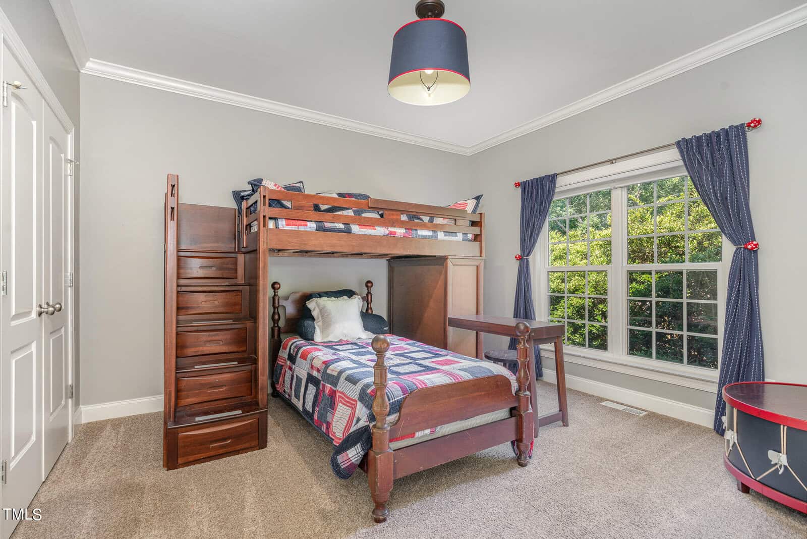 A children's bedroom with a wooden loft bed and a twin bed underneath. The room has gray walls, a large window with navy curtains, and a red and white ceiling light. A laundry basket is on the floor, and the carpet is beige.