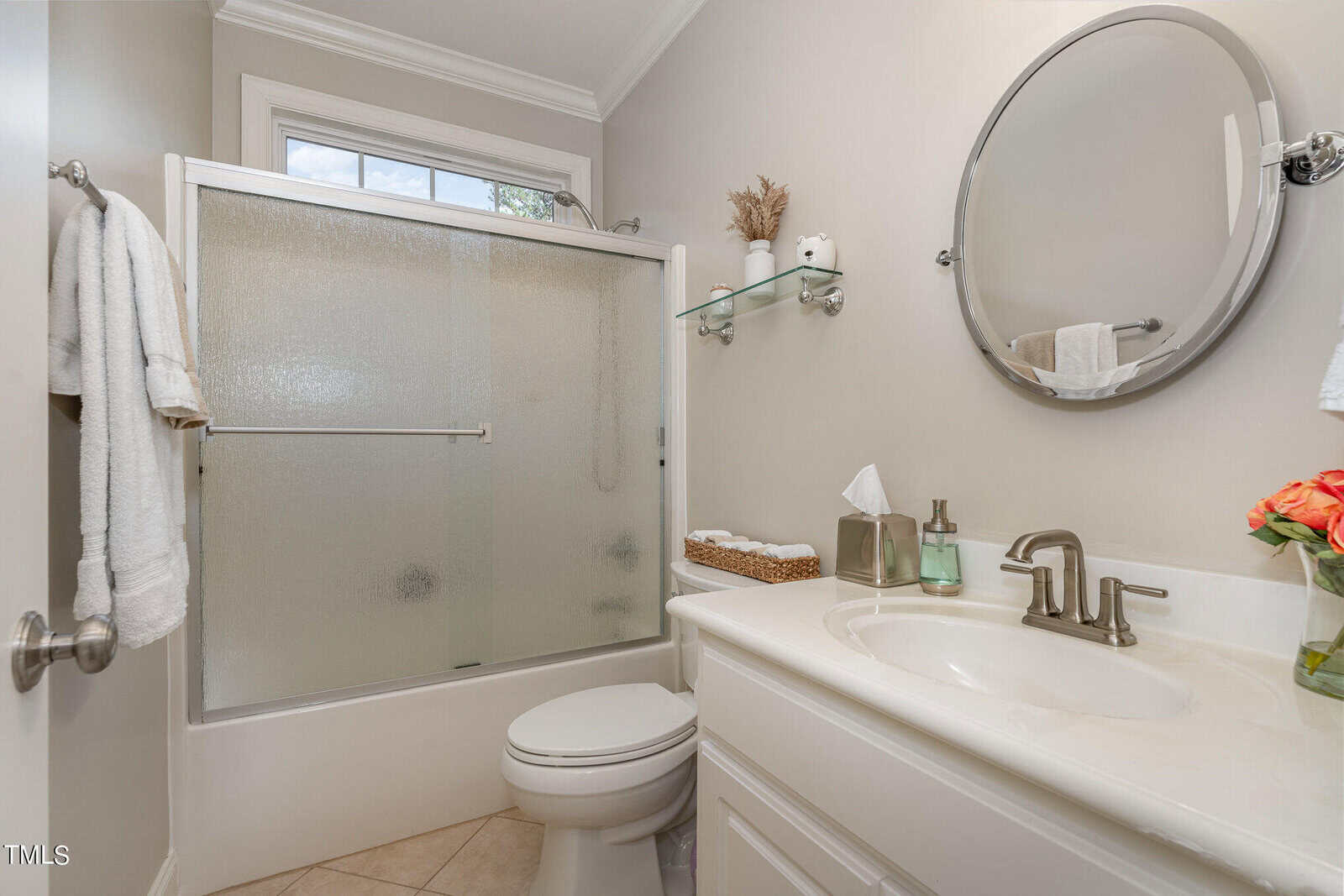 A small, well-lit bathroom with a frosted glass shower, white toilet, and a vanity with a round mirror. The sink area has a tissue box, basket of towels, and a vase of flowers. A towel hangs on a chrome bar by the door.