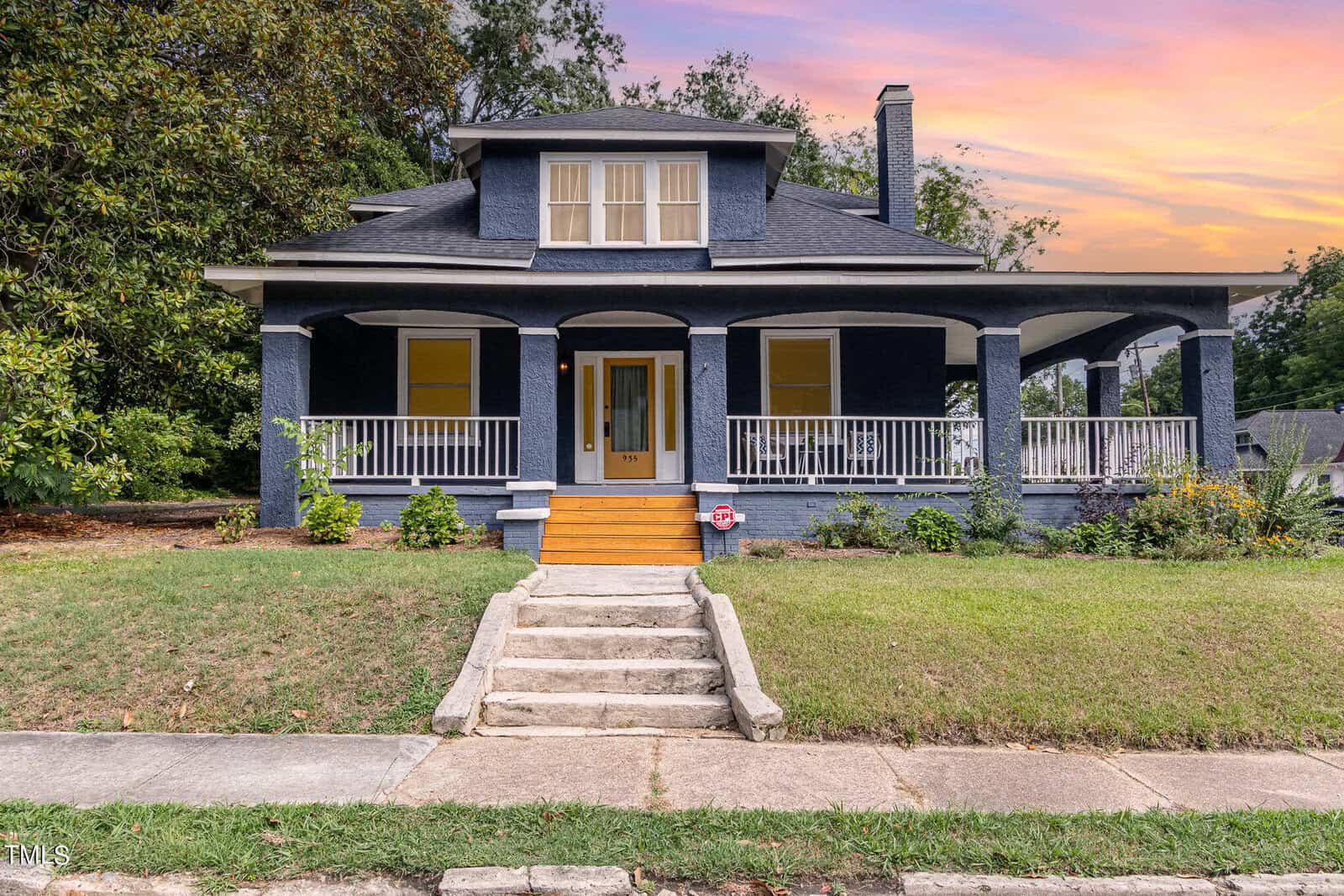 A two-story blue house with a large front porch, white railings, and wide steps leading up to the front door. Located at 935 S Chestnut Street, Henderson NC, the house is surrounded by a green lawn and various shrubs, set against trees and a colorful sunset sky.