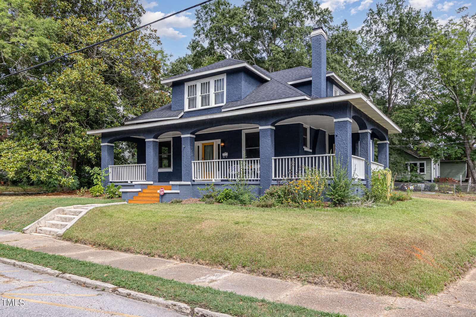 An image of a two-story house painted in dark blue with white trim. It features a large wraparound porch with white railings, a steep roof, and a prominent chimney. The house is surrounded by a well-maintained lawn and various trees and shrubs.