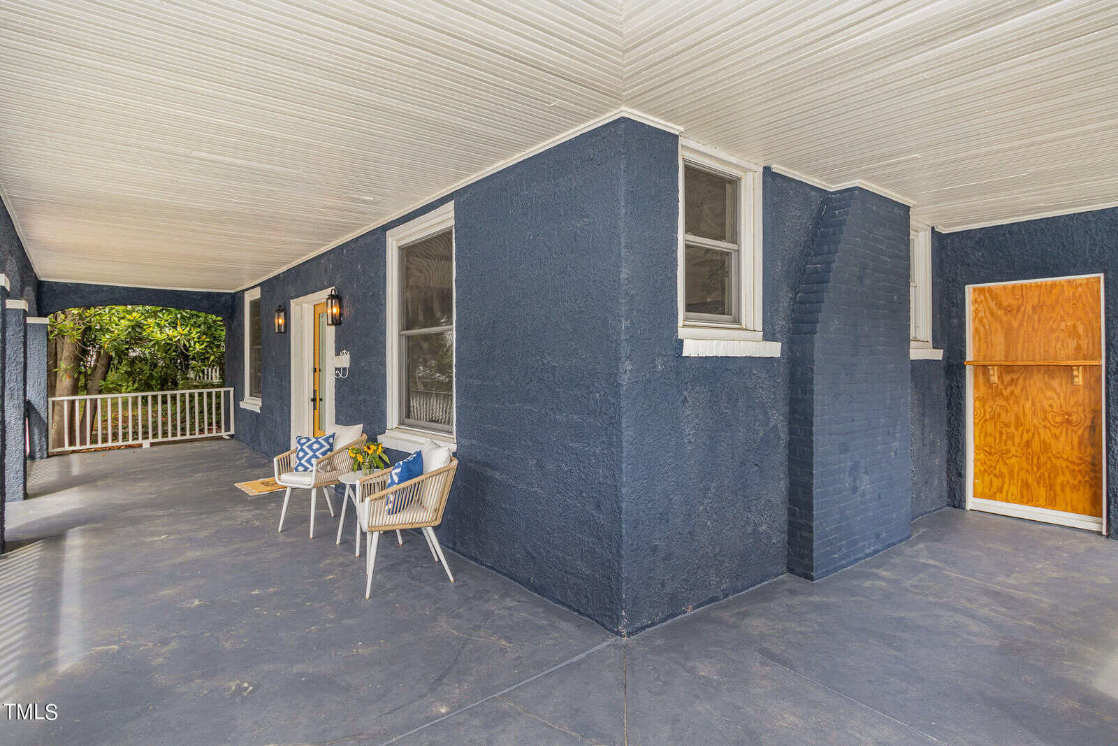 A covered porch area with dark blue walls and light-colored ceiling. The porch features a small sitting area with two chairs, a round table, and a potted plant. There are two wall-mounted lanterns by the front door. The area has a wooden board covering a section to the right.