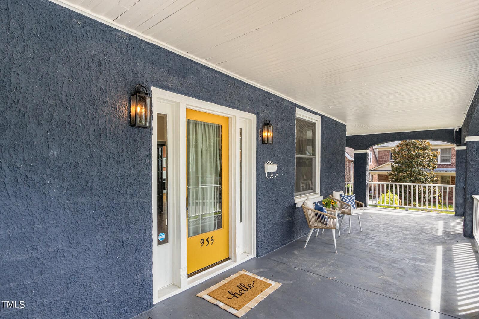 A spacious porch with a navy blue wall features a bright yellow door with the number "935" on it. Next to the door is a welcome mat with the word "hello." Two outdoor chairs with cushions are positioned on the right side, with part of a house visible in the background.