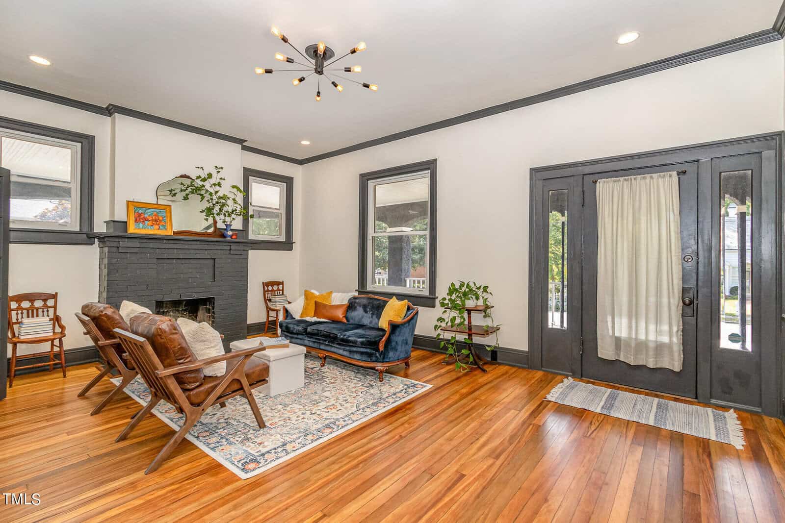 A bright living room with hardwood floors, a black door, and gray trim. It features a modern black and gold chandelier, a gray brick fireplace, wooden chairs, a blue velvet sofa with yellow cushions, and plants. A patterned rug sits under the seating area.