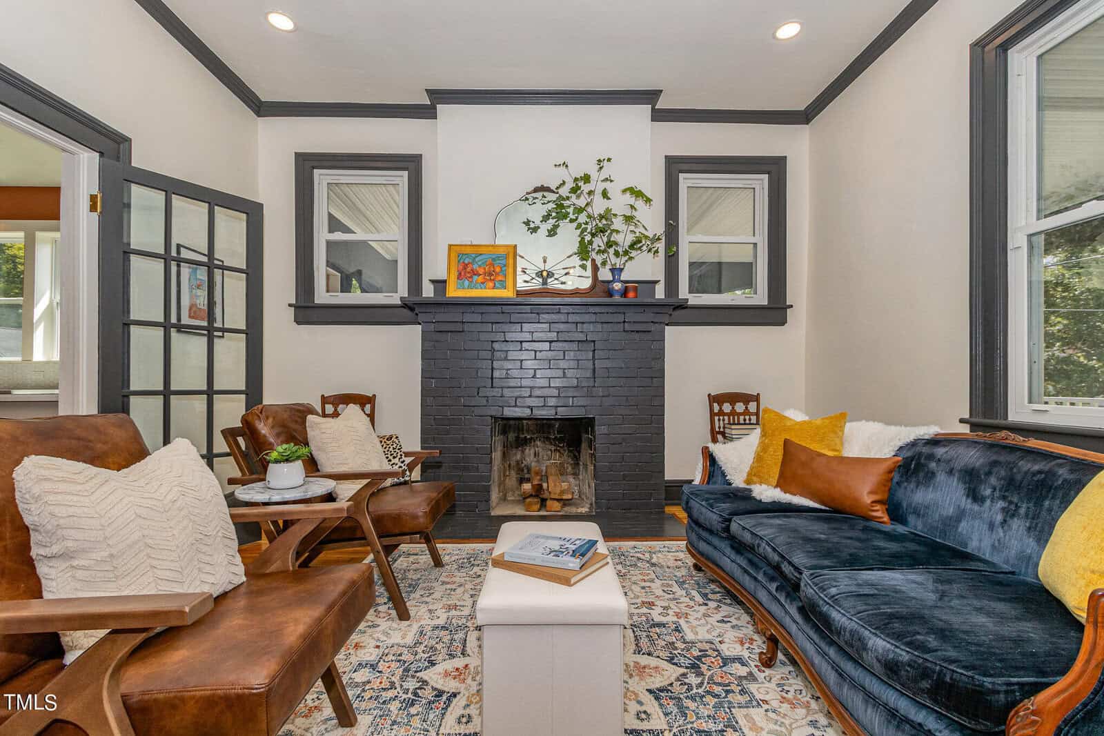 A cozy living room decorated with a blend of modern and vintage elements. It features a dark brick fireplace centered between two windows, a navy velvet couch adorned with yellow pillows, leather chairs with white cushions, and a patterned rug on the floor.