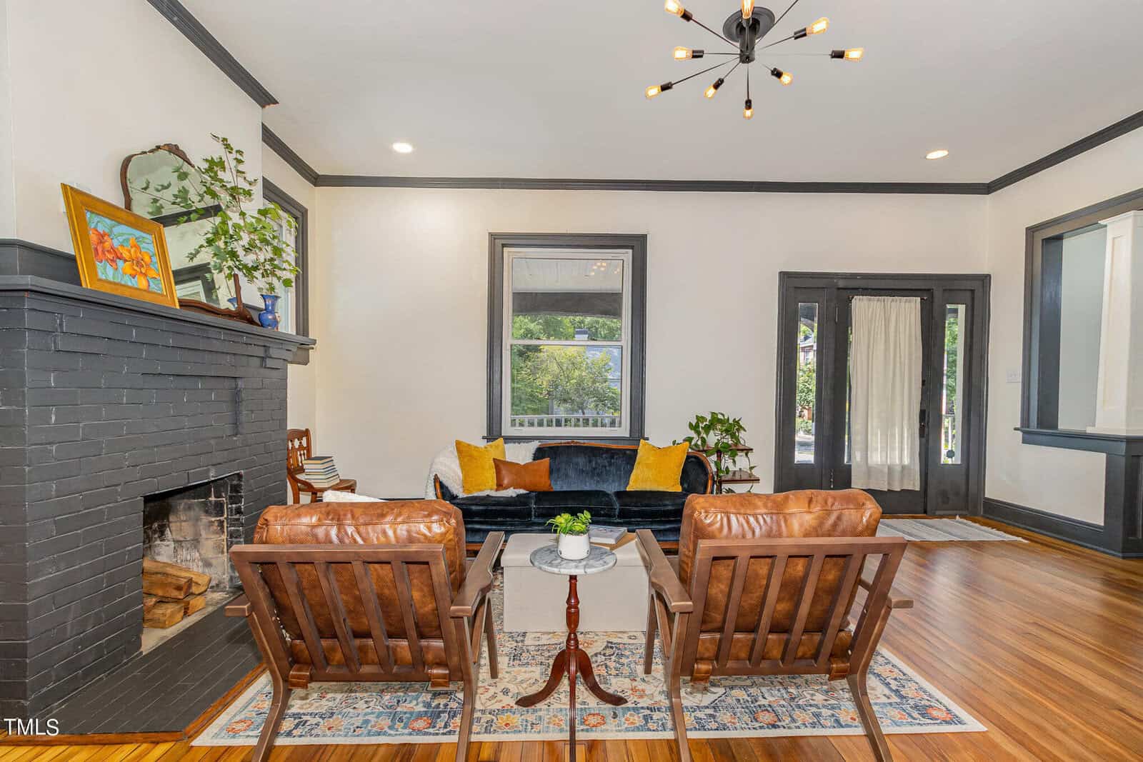 A cozy living room featuring a modern black fireplace, a dark blue velvet sofa, two brown leather chairs, and a round coffee table. The room has hardwood floors, a colorful area rug, large windows, and a unique chandelier hanging from the ceiling.