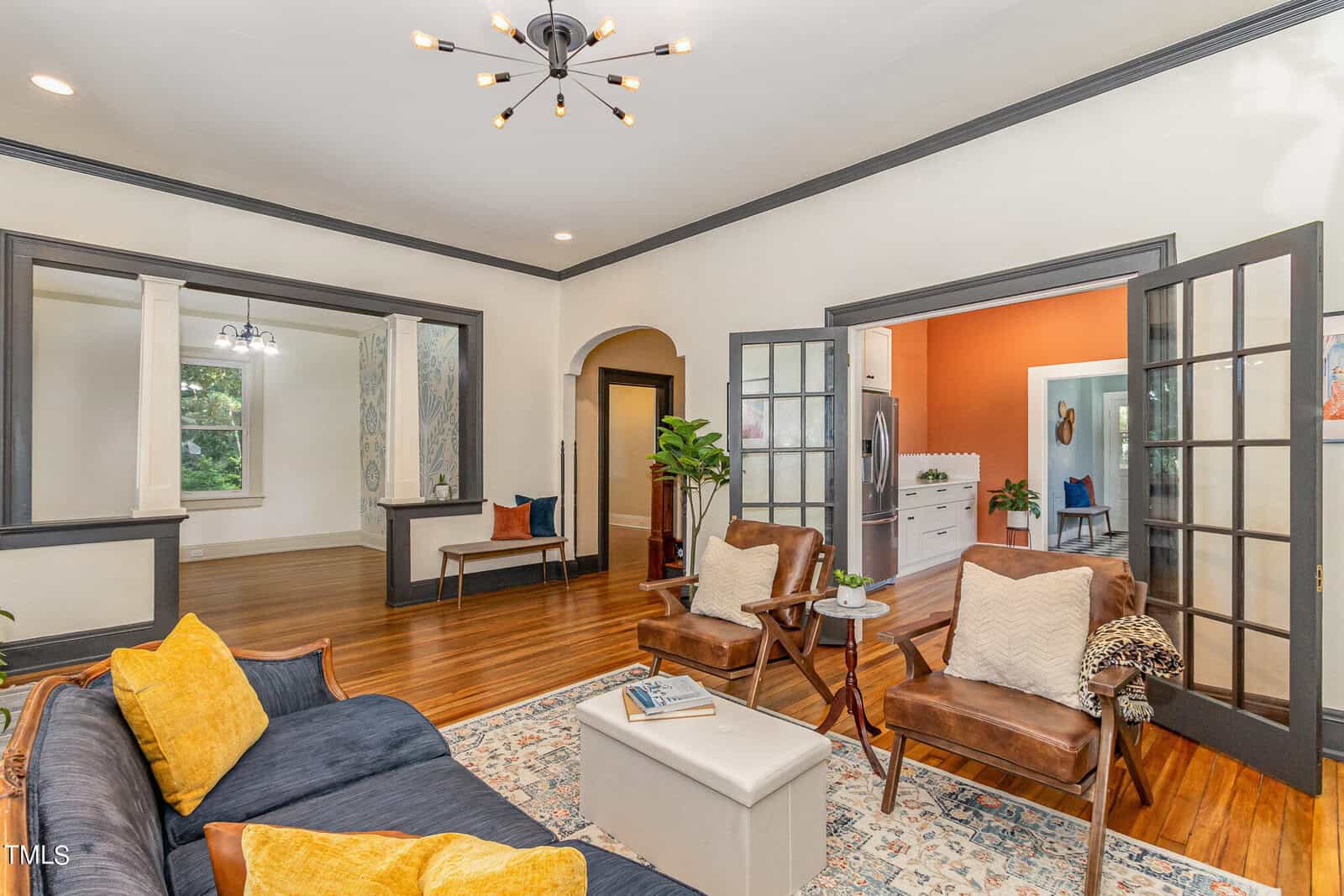 A cozy living room with wooden floors and a mix of mid-century and contemporary decor. It features a dark blue sofa with yellow pillows, two brown leather chairs, a white coffee table, and a modern chandelier. The space opens to other rooms through archways and glass doors.
