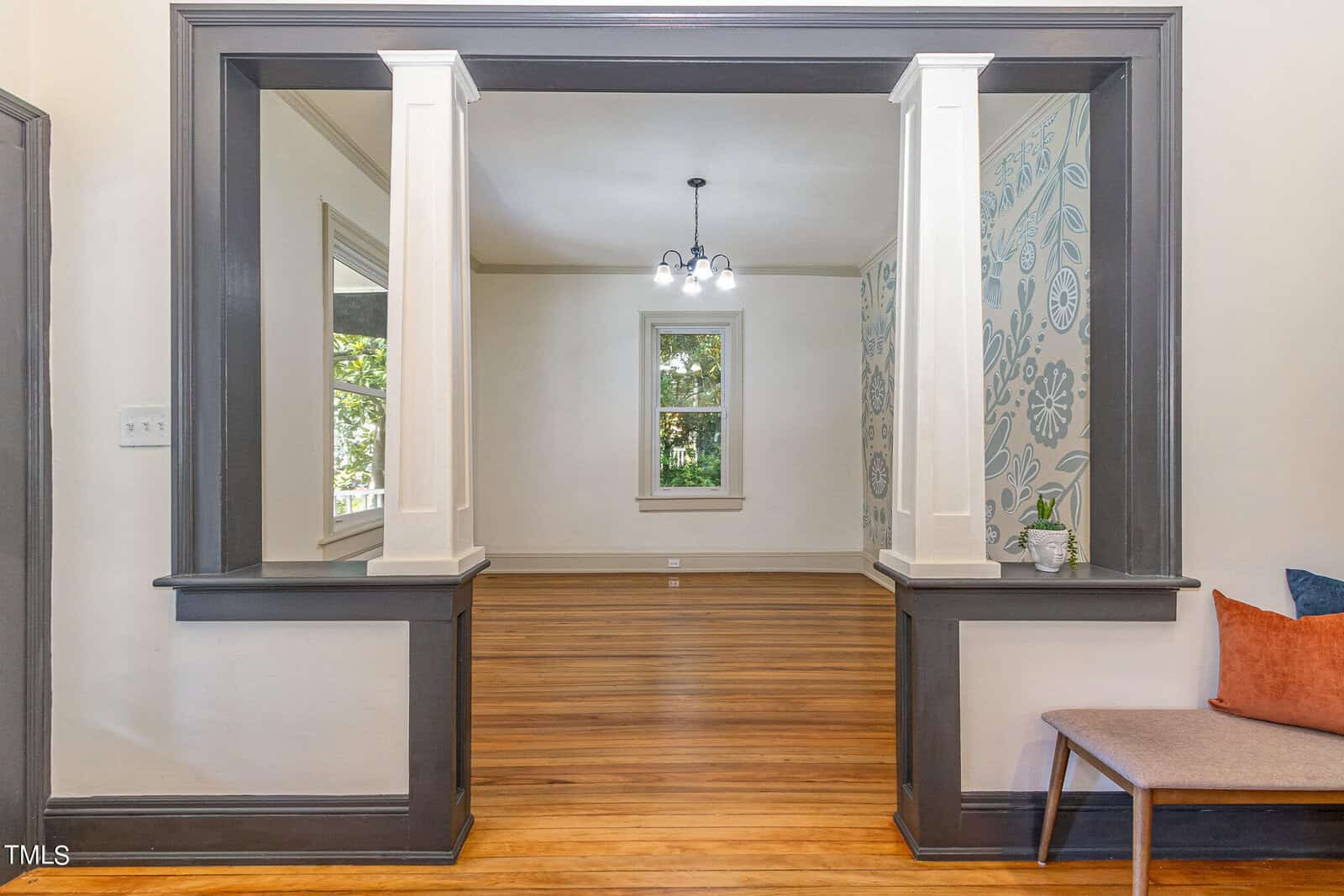 A bright, empty room with wooden floors and white walls, viewed from an adjacent room through a wide, framed opening. The far wall features floral-patterned wallpaper, and there is a single window centered on it. A modern light fixture hangs from the ceiling.