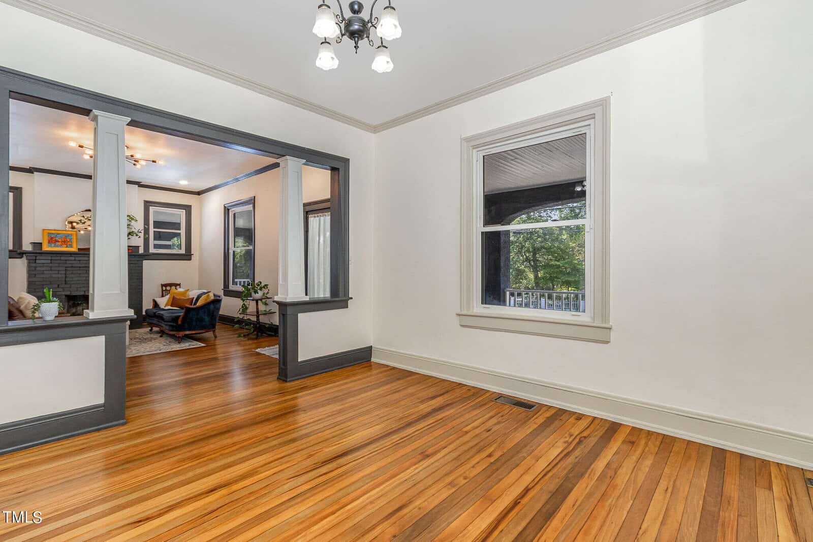 A bright room with polished wooden floors and white walls featuring grey trim. The room has a window on the right and an entryway with columns and a cutout wall to a cozy living area with a fireplace and armchairs. A light fixture hangs from the ceiling.