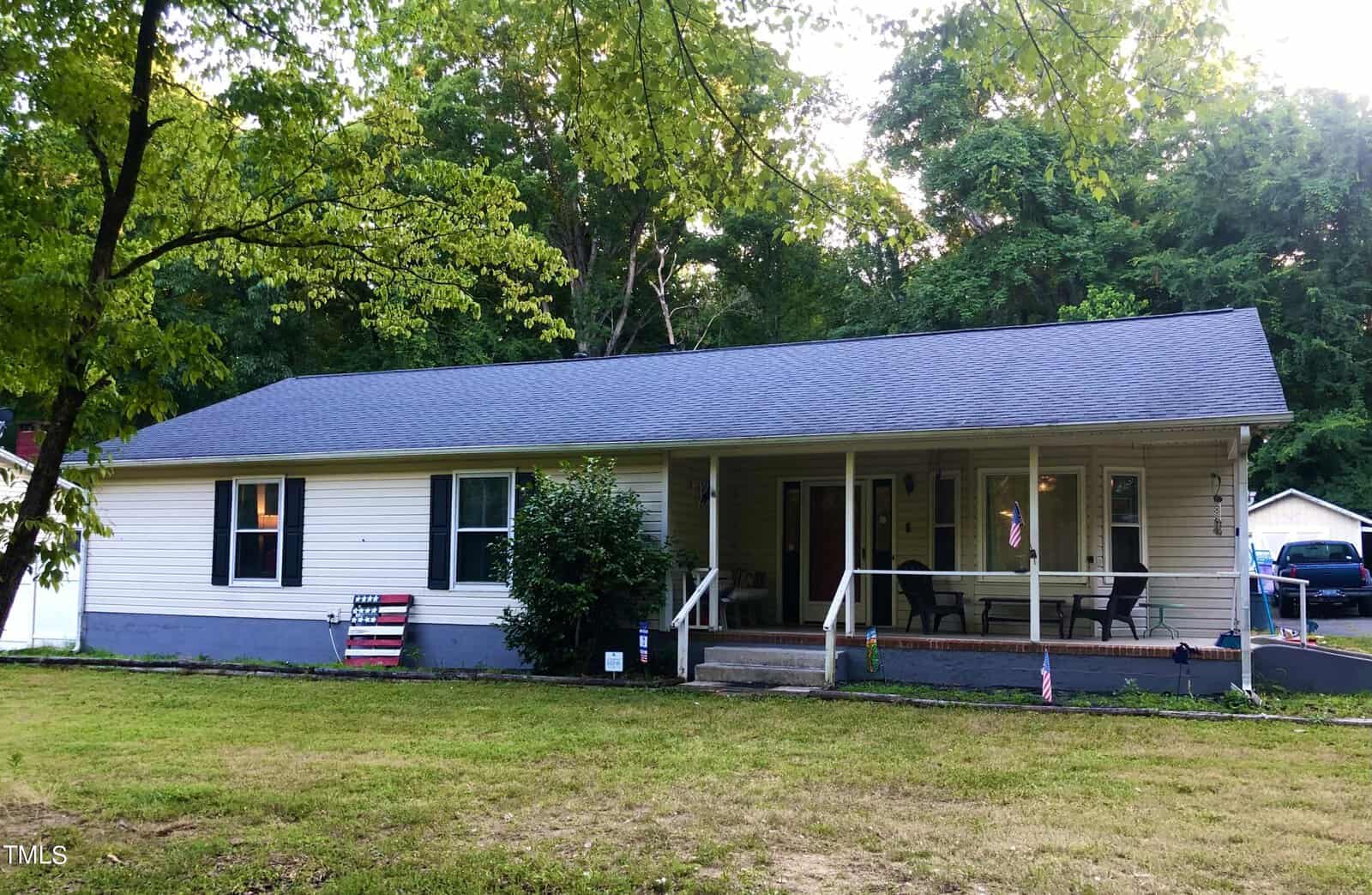 A single-story house at 1618 W B Street, Butner, features light-colored siding and a dark gray roof, nestled amid lush greenery. The cozy porch with chairs and an American flag graces the yard, while a detached garage stands proudly in the background.