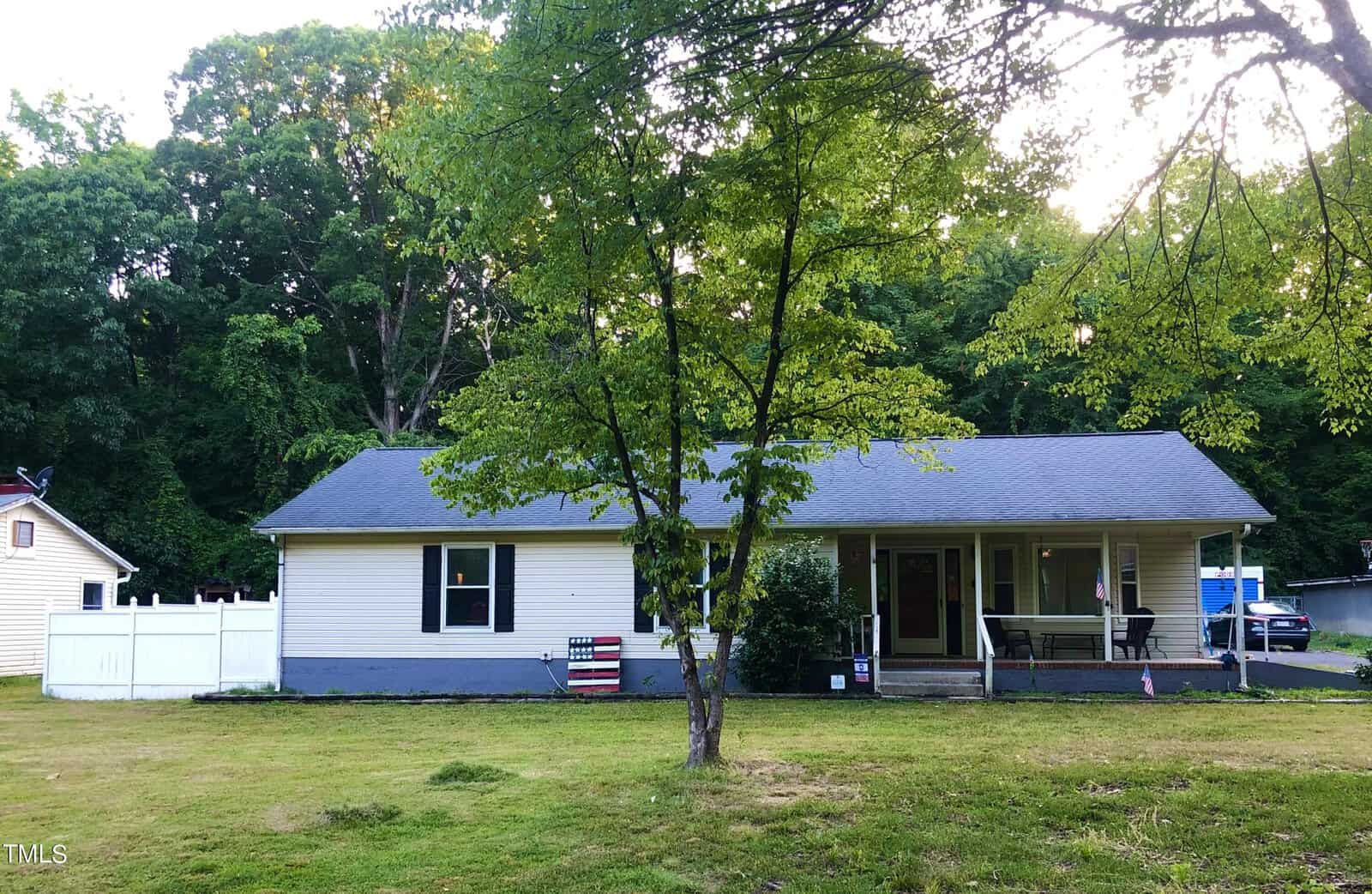 A single-story house with a light-colored exterior and dark roof sits amidst green trees. A small porch is visible, and a white fence stands to one side. The lawn is spacious with a tree in the foreground. A decorative American flag is in the yard.