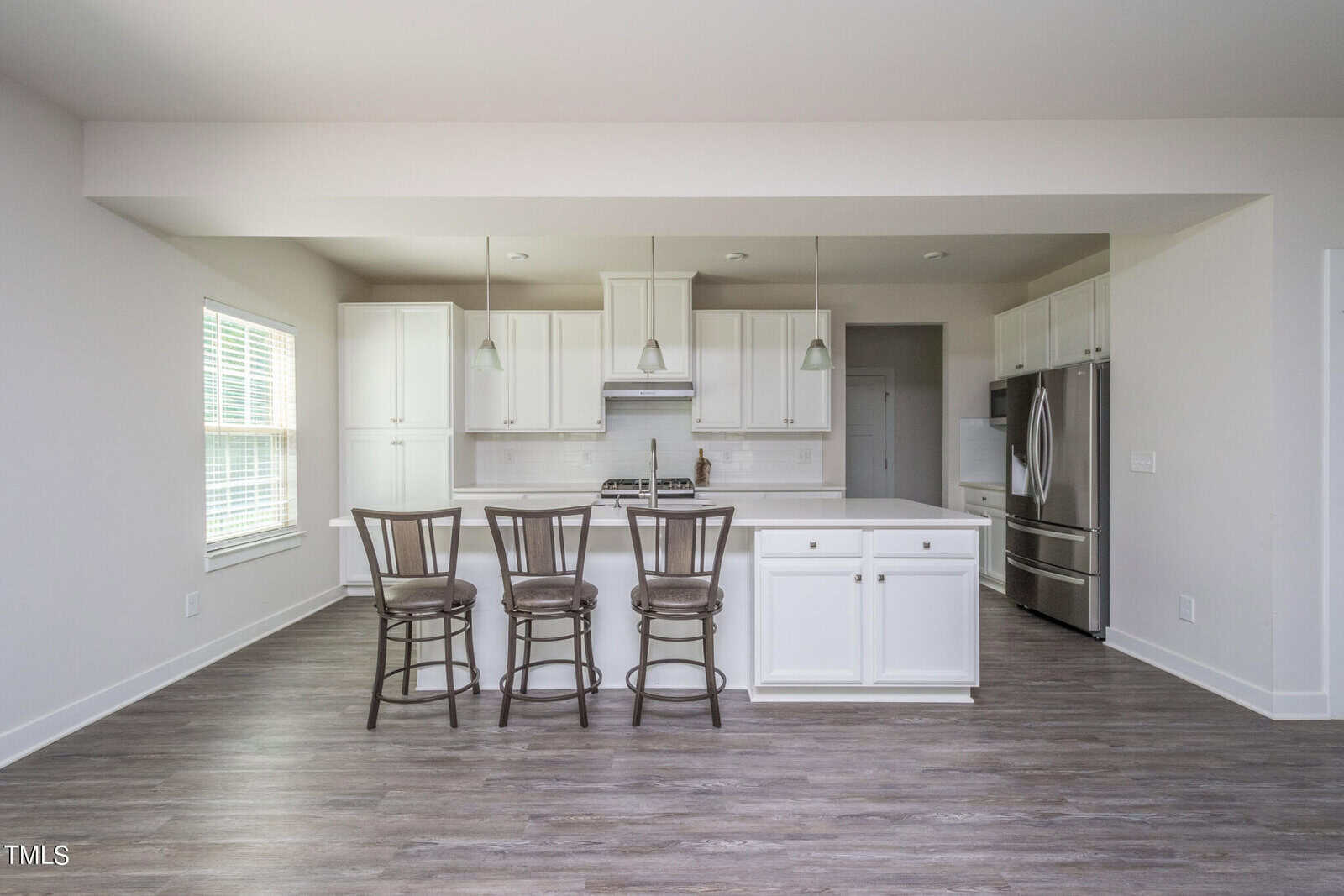 Modern kitchen with white cabinets, stainless steel appliances, a center island with three wooden barstools, pendant lights above, and wood-style flooring. Large window on the left lets in natural light.