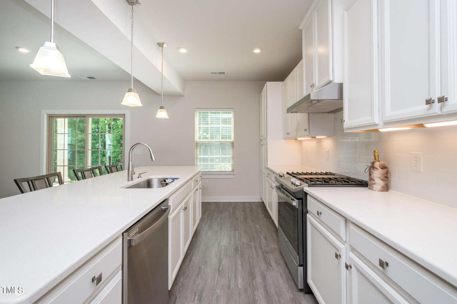 Bright, modern kitchen featuring white cabinets, stainless steel appliances, a large island with a sink, pendant lighting, and wood-look flooring. A dining area with large windows is visible in the background.