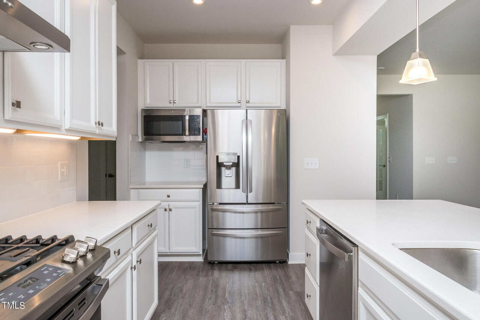 Modern kitchen with white cabinets, stainless steel appliances including a refrigerator, microwave, and oven, gray wood flooring, and white countertops; a pendant light hangs above the island sink.