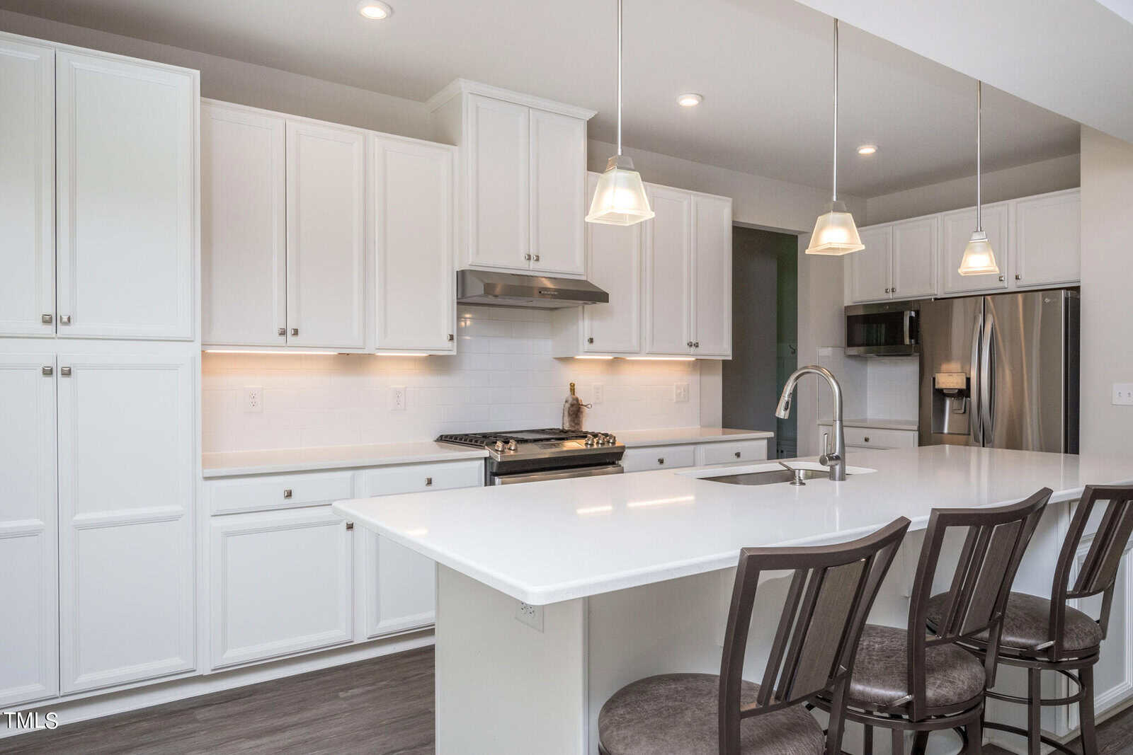 A modern white kitchen features a large island with three chairs, stainless steel appliances, pendant lights, and white cabinets with silver hardware.