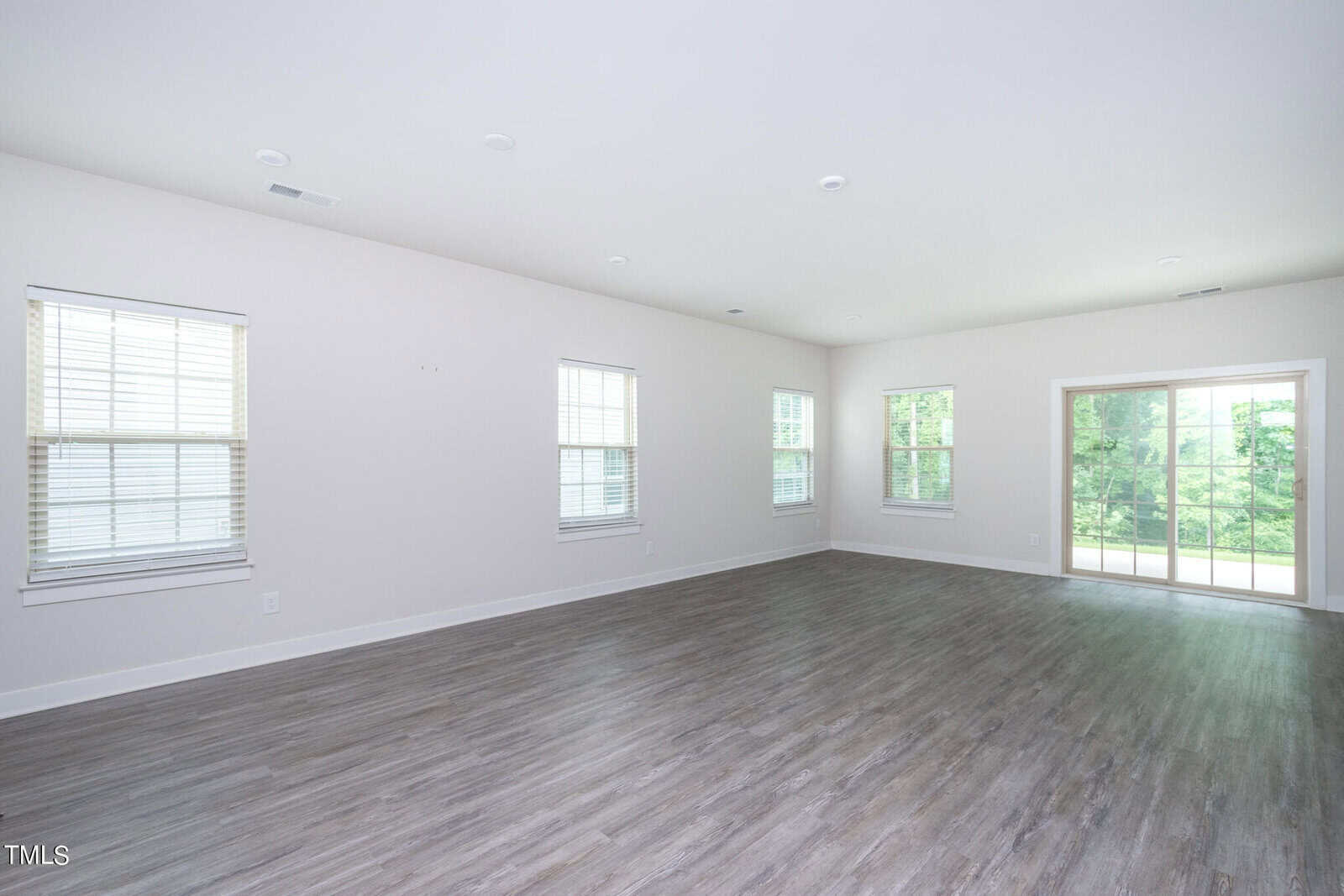 A spacious, empty room with light-colored walls, multiple windows, wood-style flooring, and large sliding glass doors opening to a view of green trees outside.