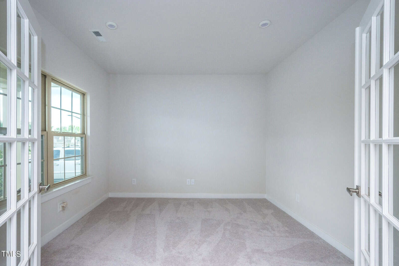 Empty room with beige carpet, light gray walls, a single window on the left, and double French doors open inward. Ceiling has two light fixtures and a vent. Floor outlets are visible near the walls.