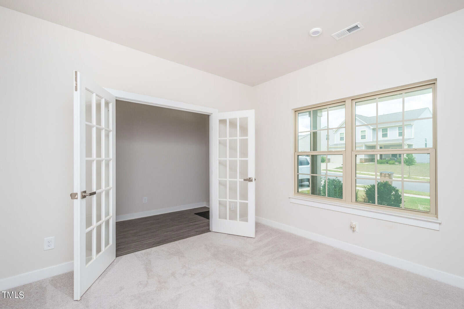A bright, empty room with beige carpet, double glass French doors, and a large window overlooking a suburban street with houses and greenery outside.