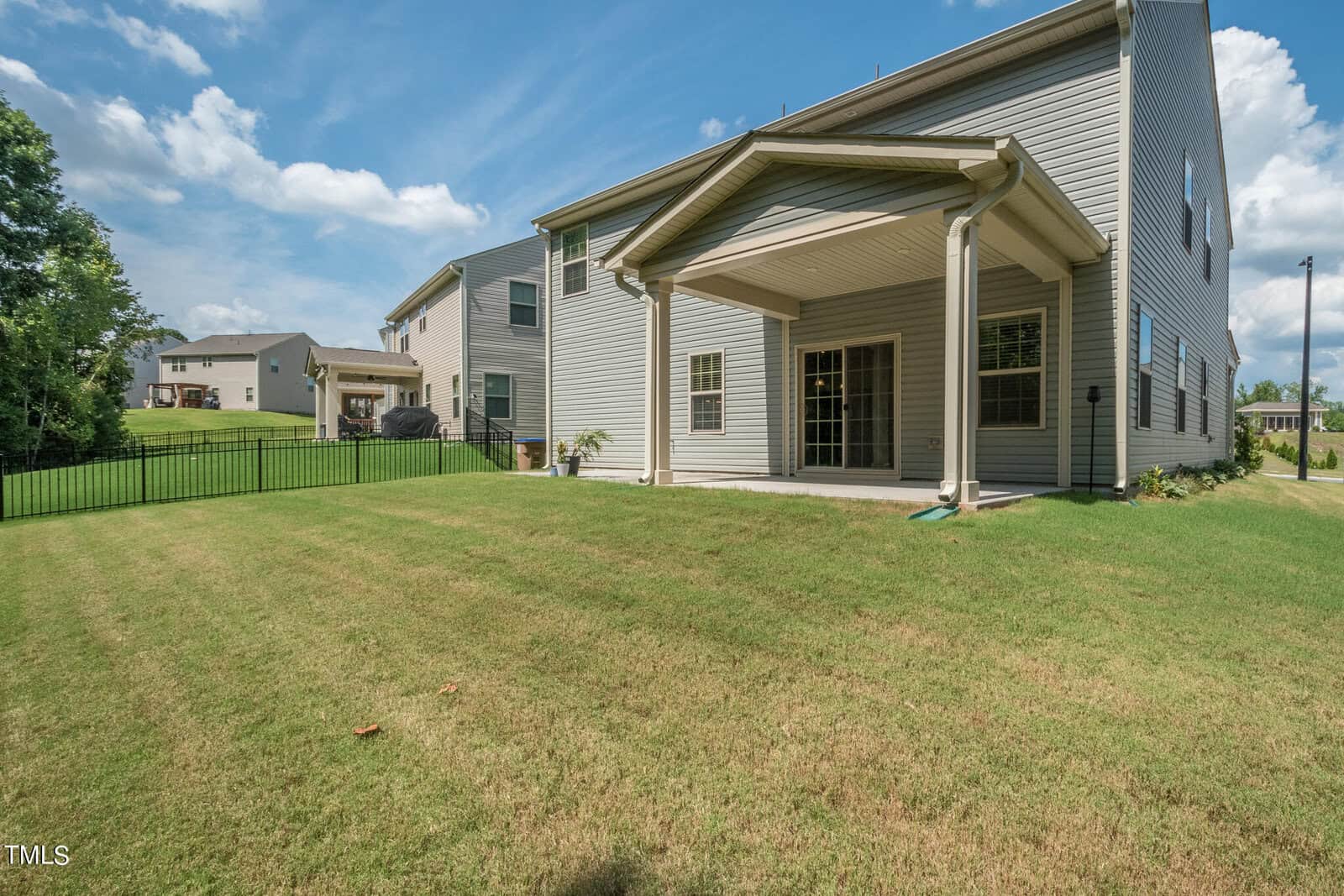 A two-story house with light-colored siding, covered back patio, and large grassy backyard, bordered by a black metal fence. Other houses and trees are visible in the background under a blue sky with clouds.