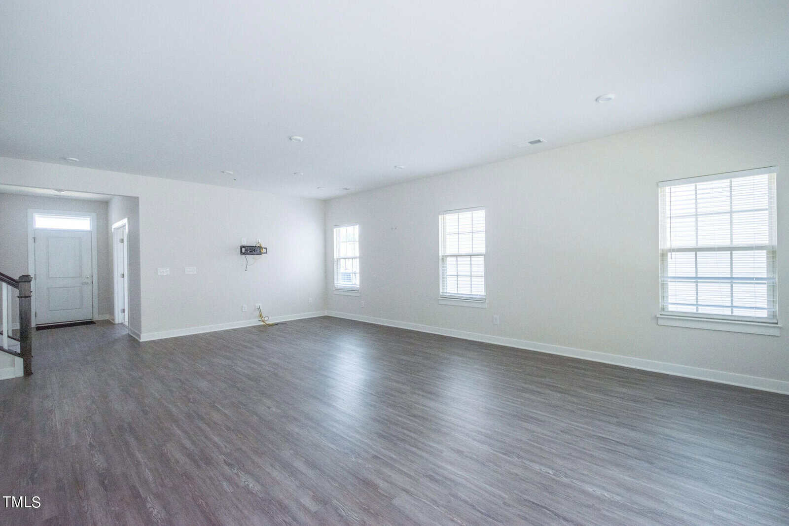 Spacious, empty living room with light gray walls, large windows, and wood-style flooring. A white front door is visible in the background, along with a staircase and a small wall-mounted fixture.