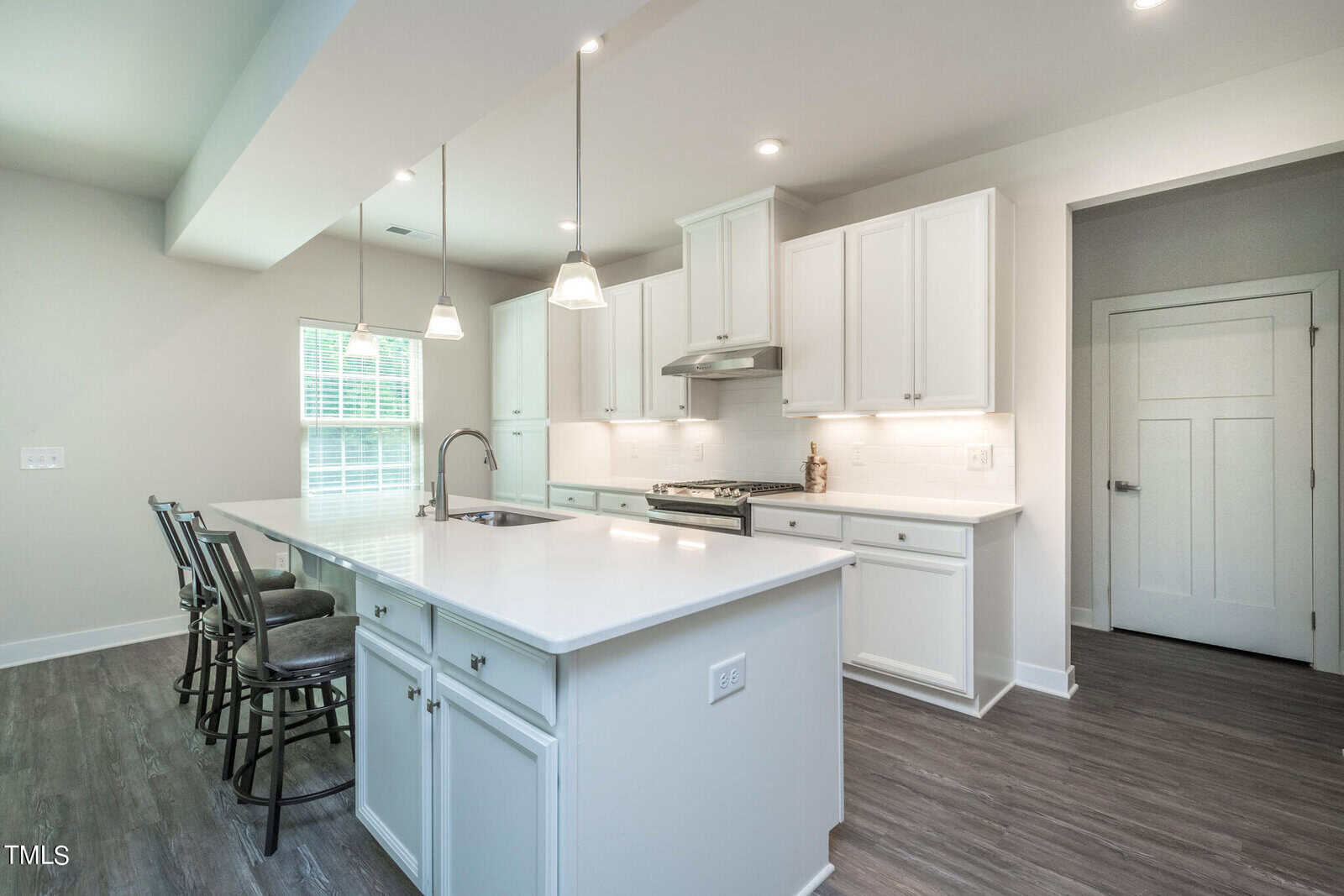 Modern kitchen with white cabinets, white countertops, stainless steel appliances, a large island with a sink, three barstools, pendant lighting, and wood-style flooring. A window lets in natural light.