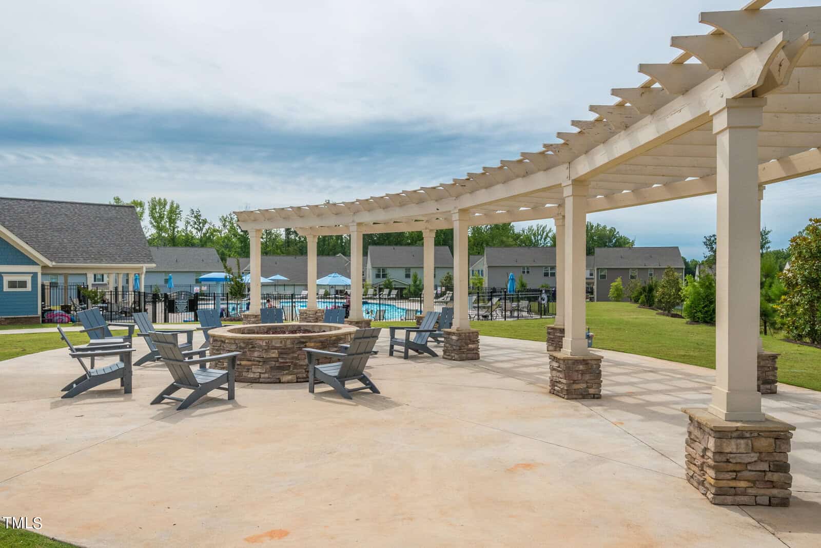 A landscaped outdoor area with a pergola, stone fire pit, and Adirondack chairs. In the background, there is a swimming pool, lounge chairs, and residential houses surrounded by green grass and trees.