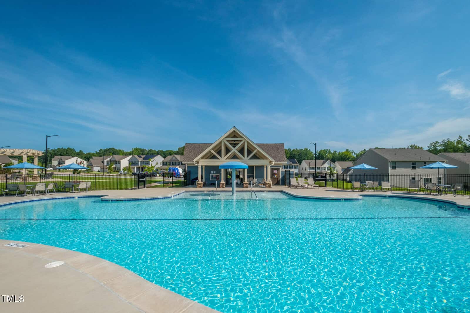 A large outdoor swimming pool with clear blue water, surrounded by lounge chairs and umbrellas, sits in front of a clubhouse with a peaked roof. Suburban houses and green lawns are visible in the background under a bright blue sky.