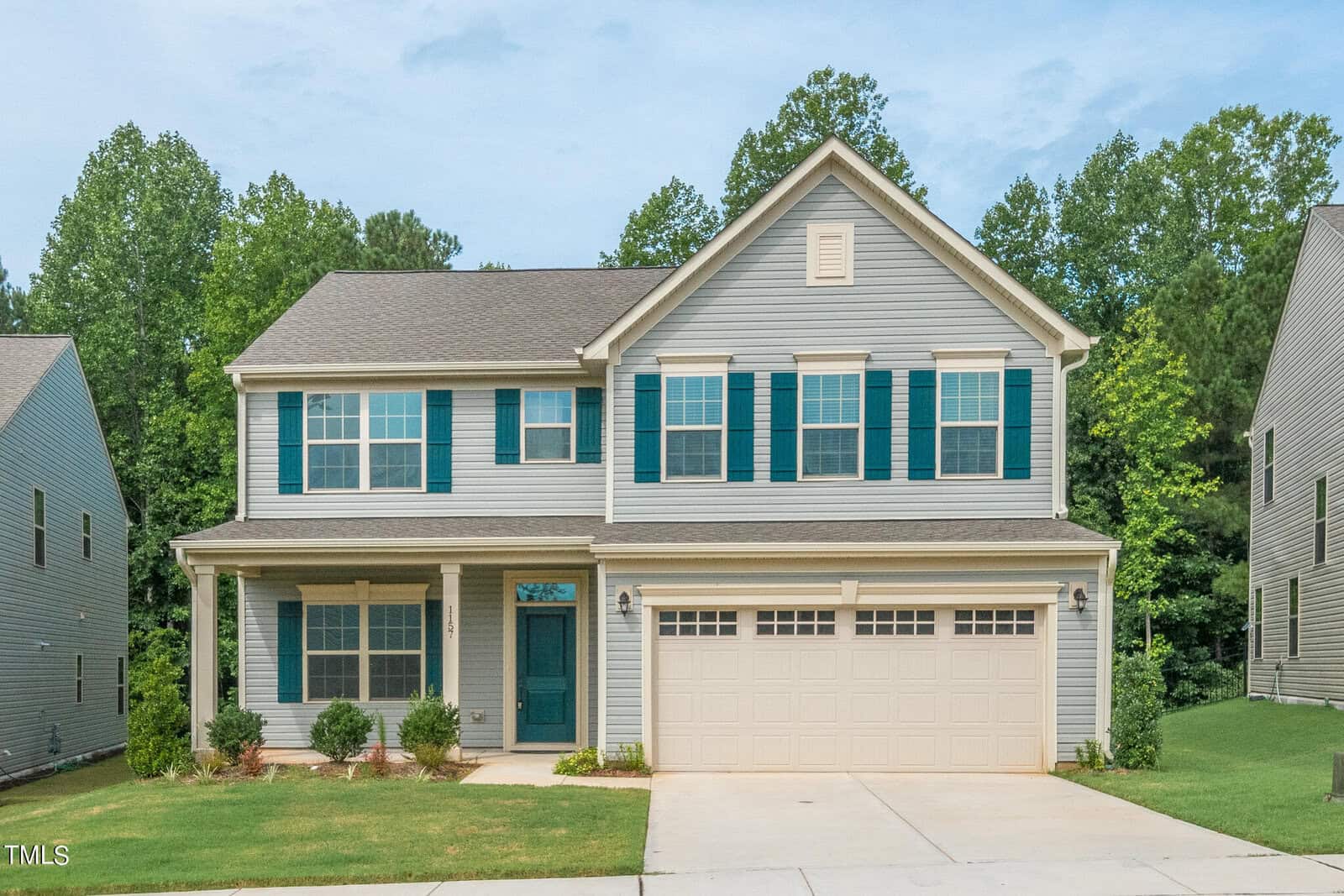 A two-story house with light gray siding, teal shutters, a covered front porch, and a double garage, surrounded by green trees and a neatly mowed lawn.