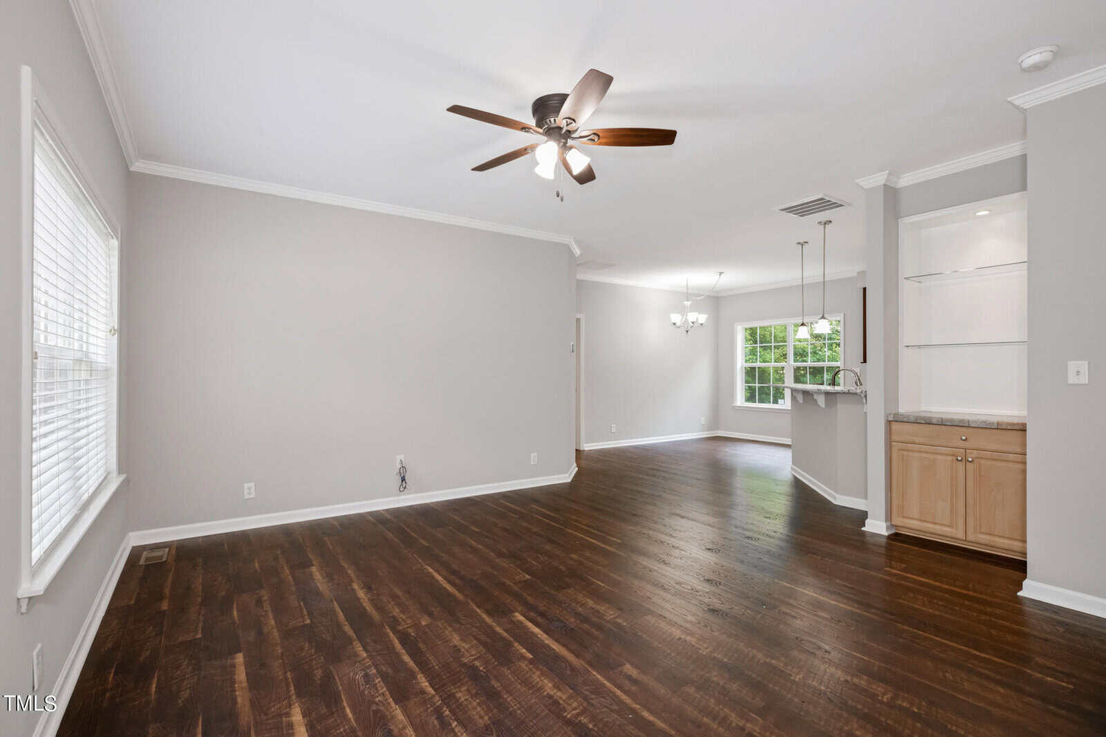 A spacious, unfurnished living area featuring dark wooden floors and light gray walls with white trim. A ceiling fan with light is mounted on the ceiling. An open floor plan connects to a dining area and a kitchen with light wood cabinetry and built-in shelving.