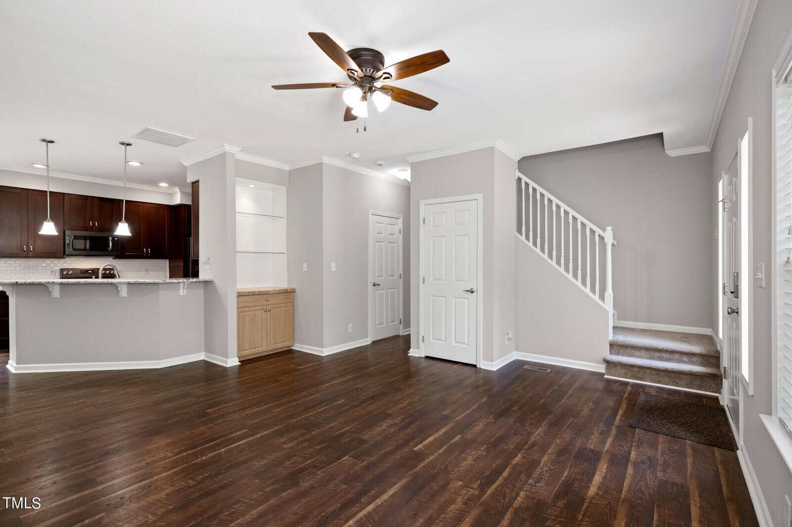 A spacious, empty living room with dark hardwood floors, light grey walls, and a ceiling fan. The room has an open floor plan leading to a kitchen with dark wood cabinets and a breakfast bar. A staircase with white railings and a small built-in cabinet are also visible.