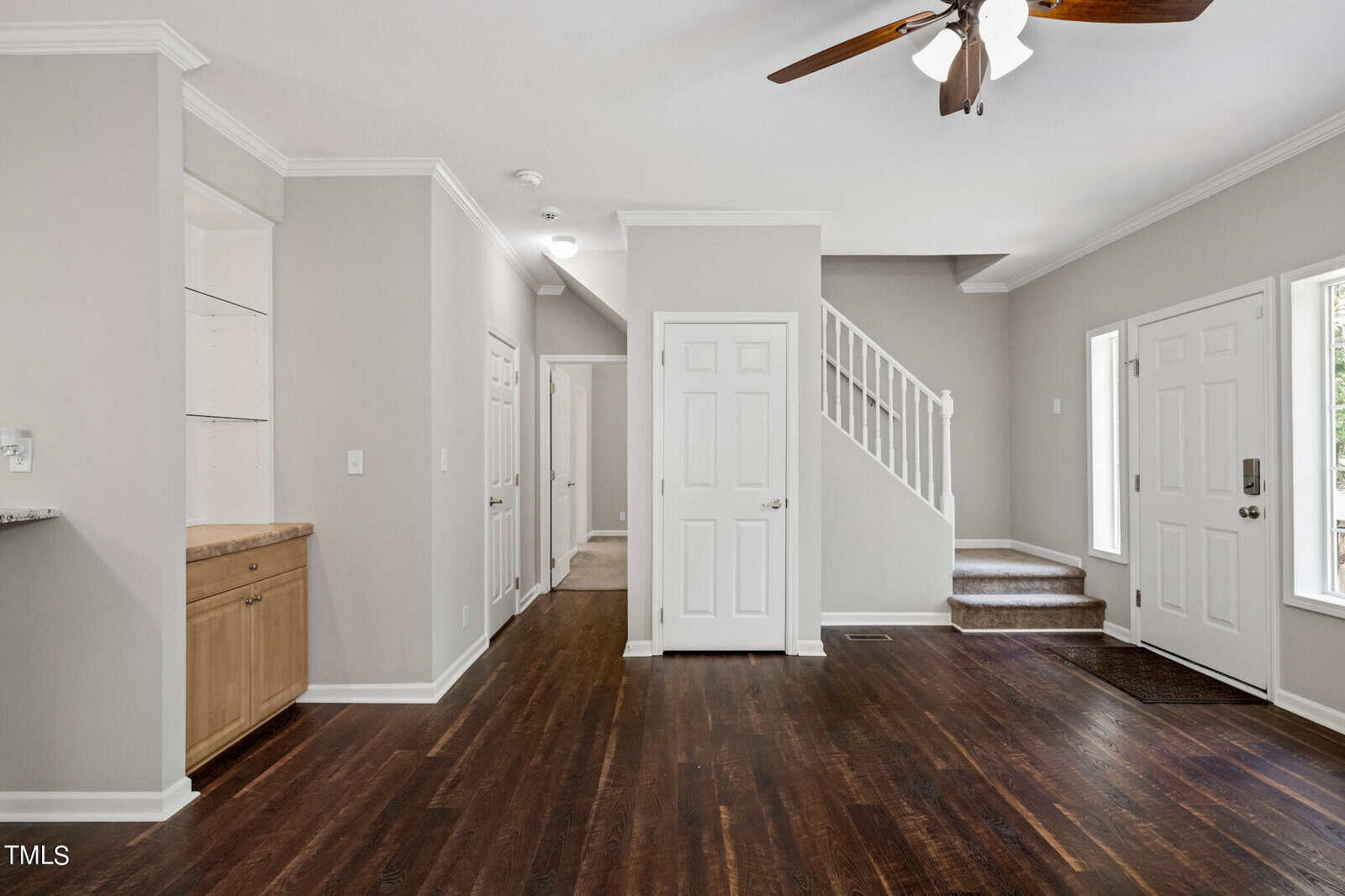 A modern interior view of a house featuring dark hardwood floors, light gray walls, and white trim. Visible are a staircase with a white railing, multiple doors, large windows providing natural light, and a built-in corner shelf with a wooden cabinet below it.