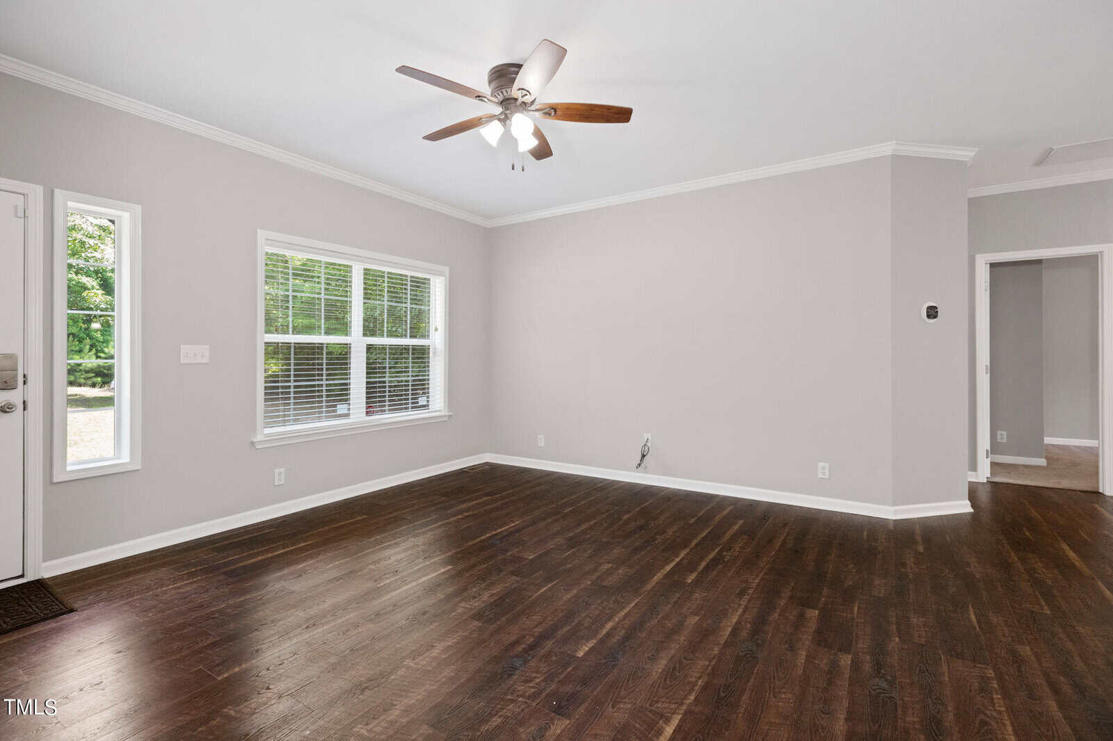 A spacious, unfurnished living room with grey walls, dark hardwood floors, a ceiling fan with a light, and a large window letting in natural light. There are doorways leading to other rooms, and a thermostat is visible on the wall.