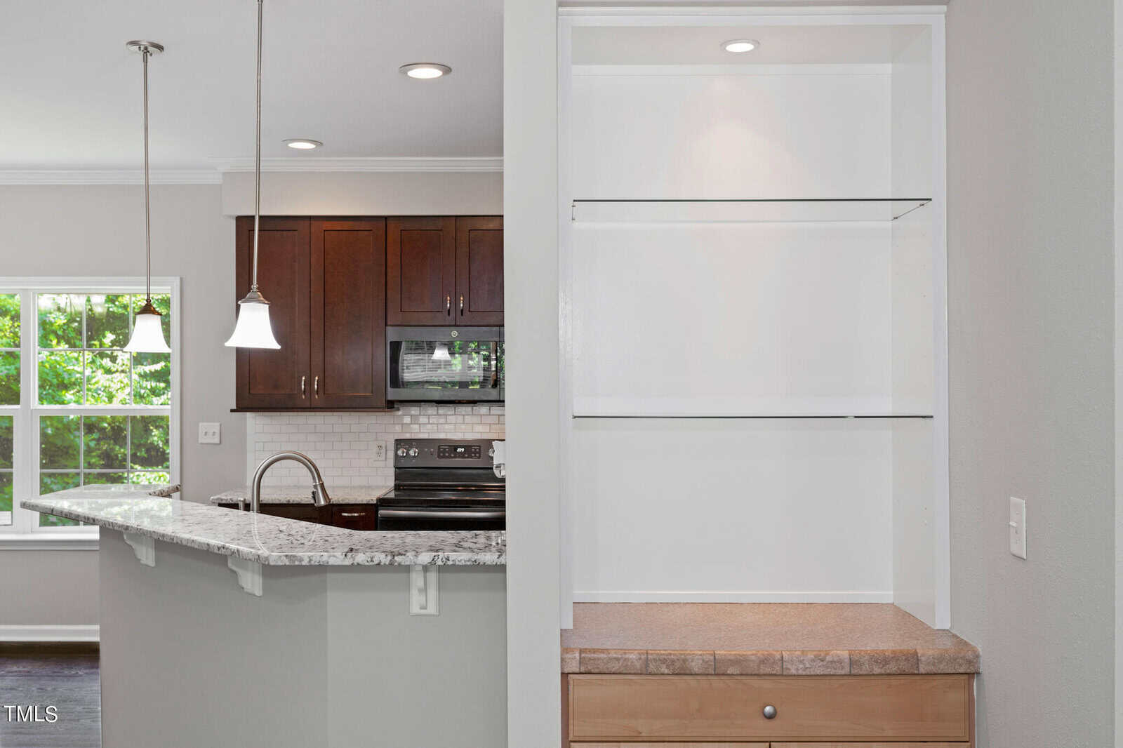 A modern kitchen with granite countertops, a breakfast bar, pendant lighting, and dark wooden cabinets. Stainless steel appliances include a microwave and electric stove. The adjacent wall features a built-in shelving unit with a light wood cabinet below.