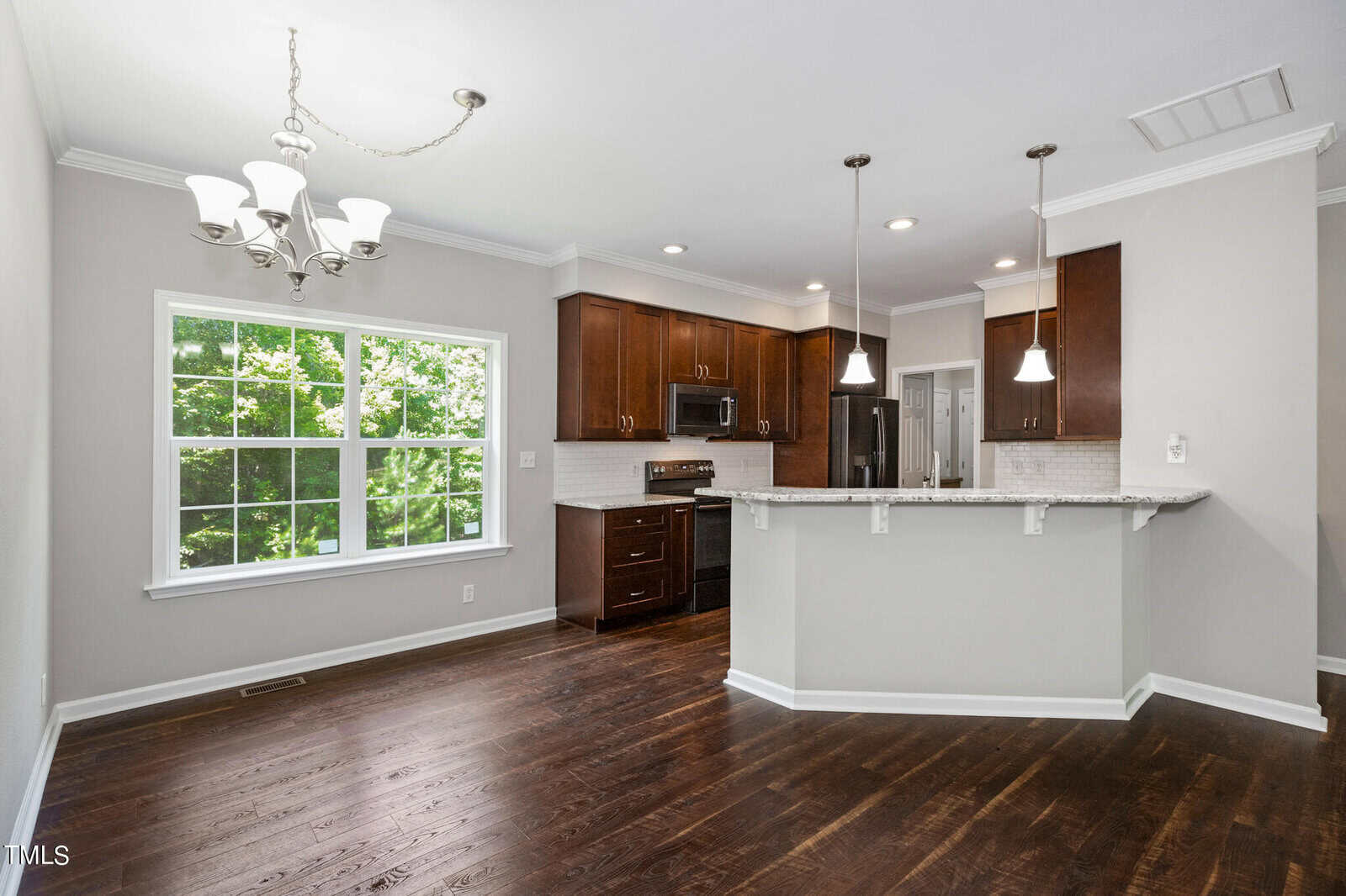 A modern kitchen and dining area with dark hardwood floors, white walls, and a large window allowing natural light. The kitchen has dark wood cabinets, stainless steel appliances, and pendant lights above a granite countertop island. A silver chandelier hangs above the dining space.