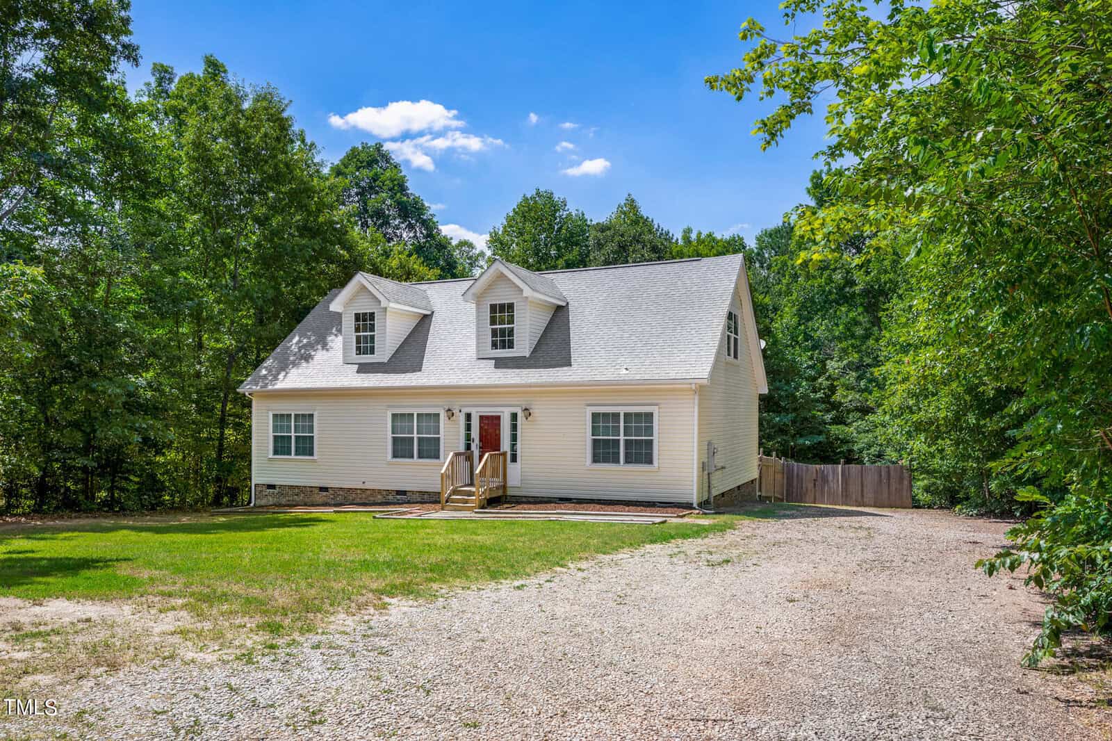 A white, two-story house with a gray roof and a red front door stands amidst a lush, green wooded area. The house has dormer windows on the second floor and a gravel driveway leading up to the entrance, which is accessible by a small set of stairs. The yard is surrounded by trees.