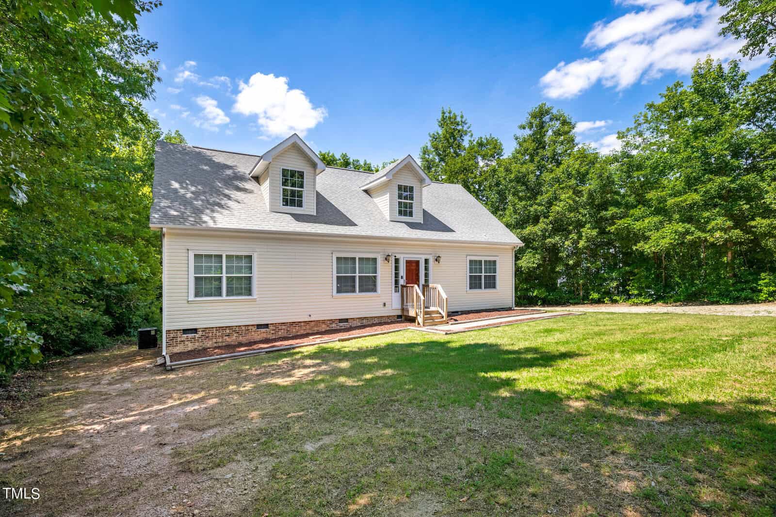 A beige two-story house with a gray shingled roof sits in a clearing surrounded by trees. The front features two dormer windows and a small porch with a wooden railing. The sky is clear and blue, and the yard is a mix of grass and dirt.
