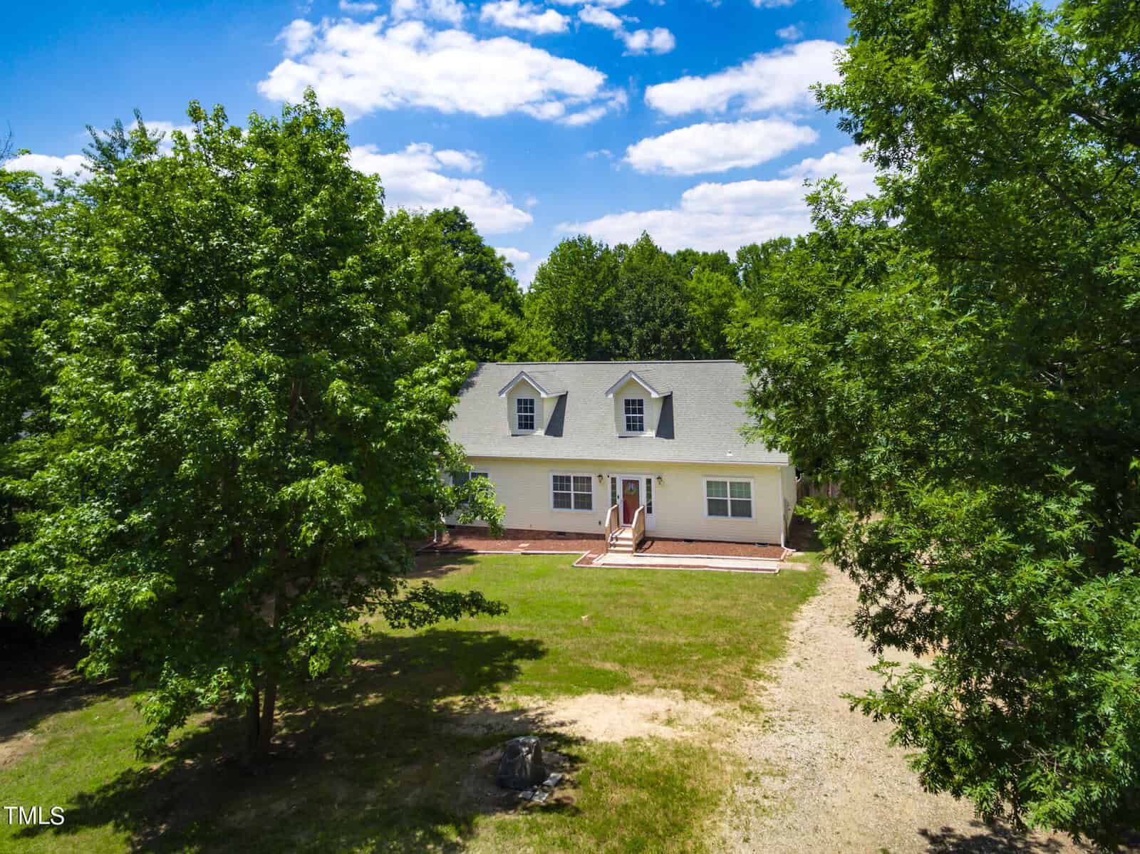 A single-story house with a gray roof, white siding, and red brick detailing, surrounded by lush green trees. A gravel driveway leads to the front steps and entrance. The property is well-maintained with a clear blue sky in the background.