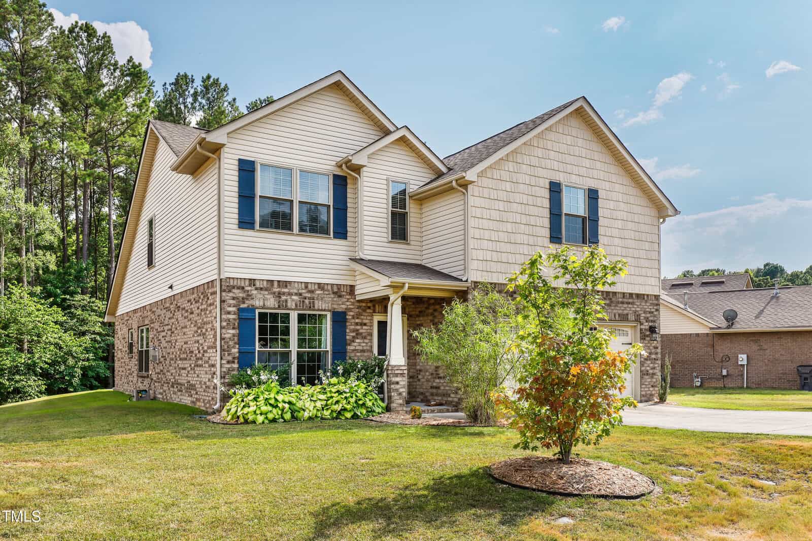 A two-story house with beige siding and brick accents, featuring blue shutters. It has a small, landscaped front yard with a tree and shrubs, and a paved driveway leading to a garage on the right. The background includes trees and a clear sky.
